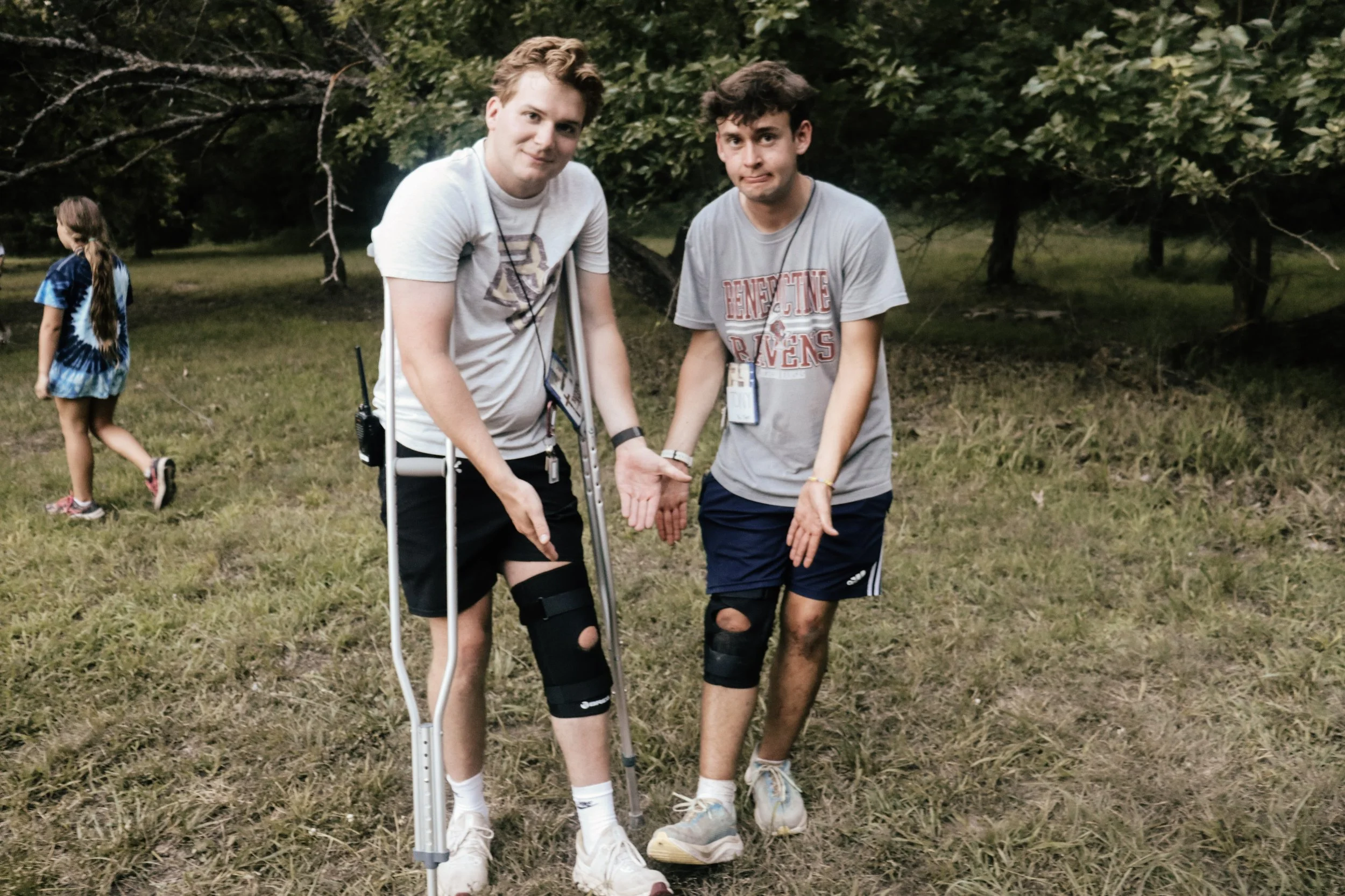 Two young men standing outdoors on grass, one with crutches and a knee brace, both with hands together, and a girl in the background wearing a tie-dye shirt walking away.