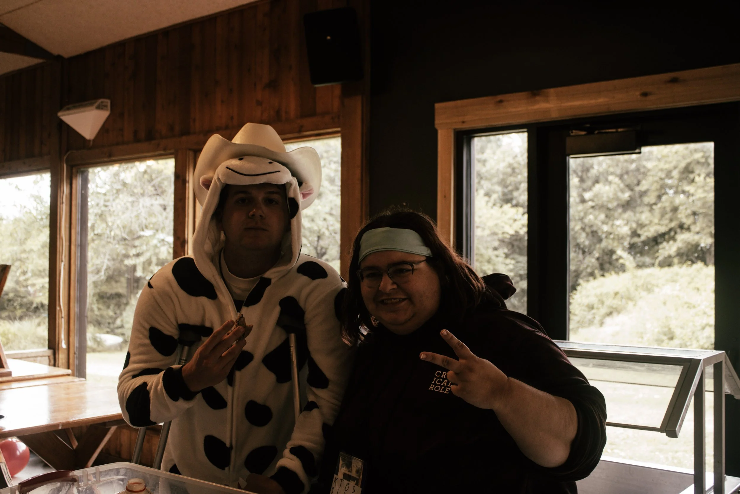 Two people posing together indoors near windows, one dressed in a cow-themed hoodie and hat, the other in a black shirt, both making peace signs.