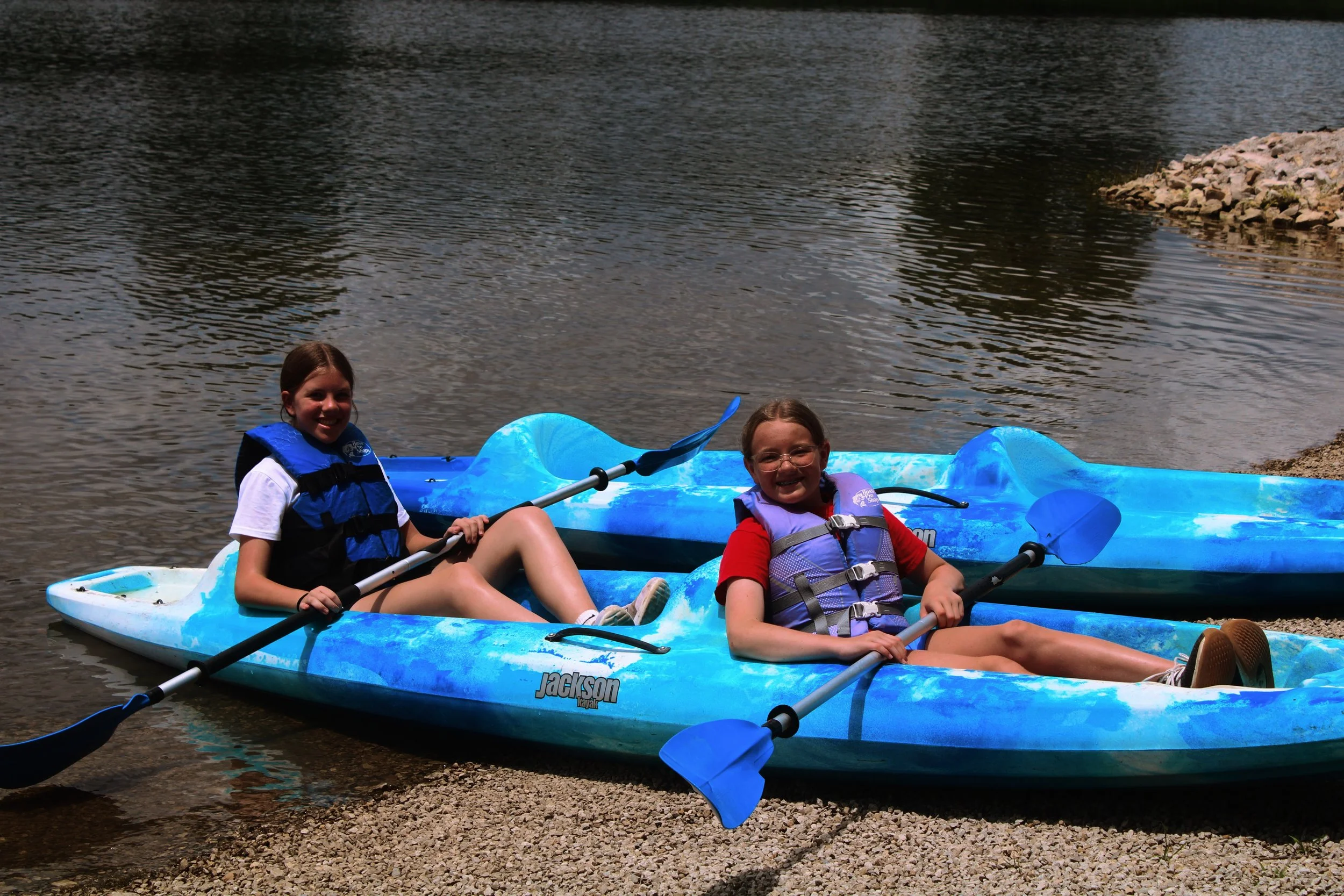 Two girls relaxing in blue kayaks on the shore of a lake, smiling, wearing life jackets, with water and rocks in the background.
