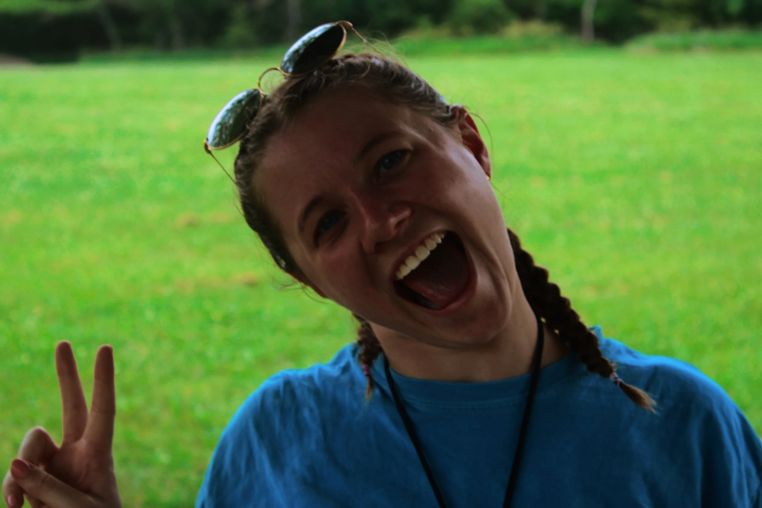 A young woman with braided hair and sunglasses on her head, smiling widely, showing her teeth, outdoors on a grassy field. She is making a peace sign with her left hand and wearing a blue shirt.