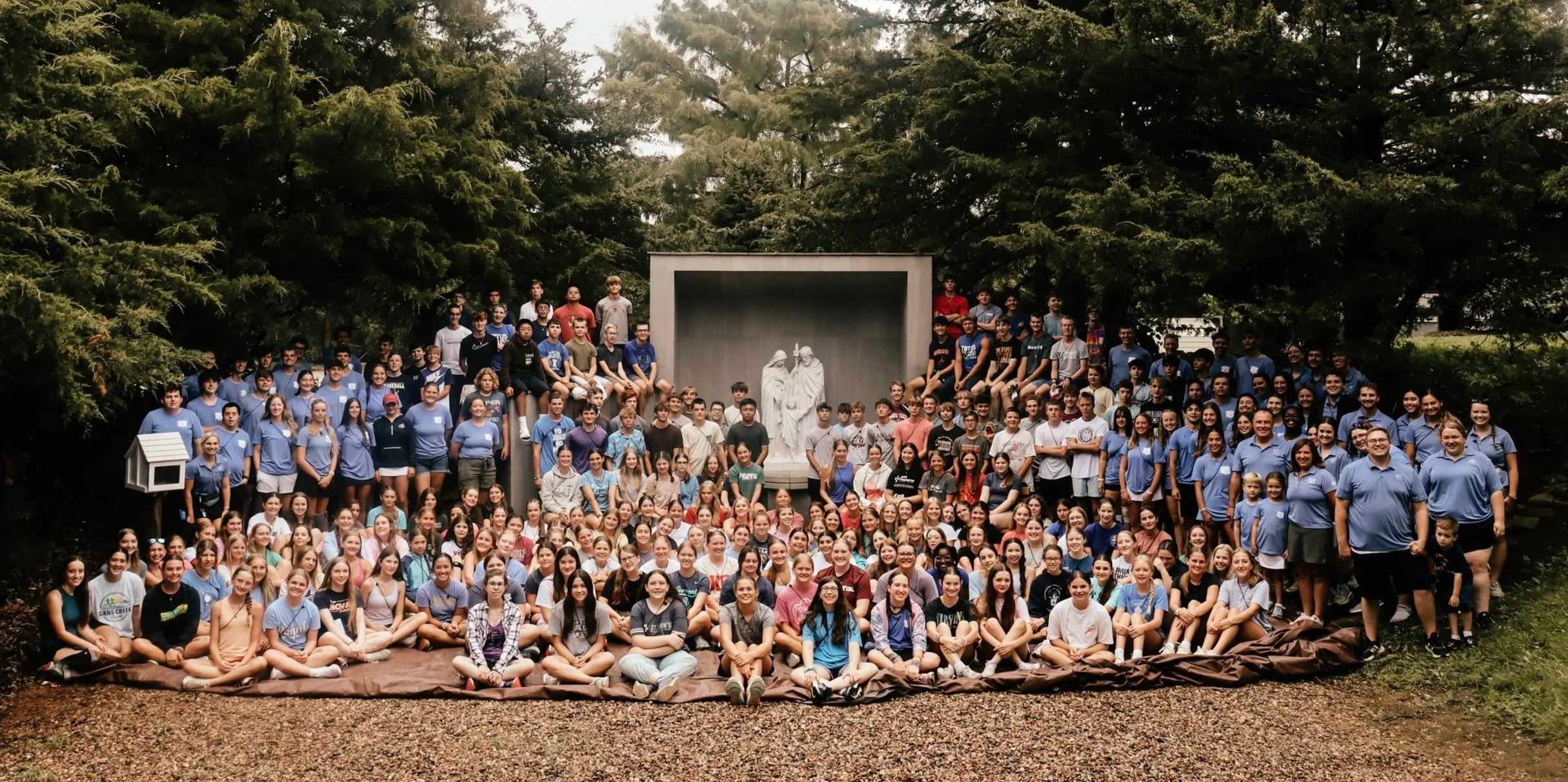 A large group of students and teachers gathered outdoors for a photo in front of a statue with trees in the background.