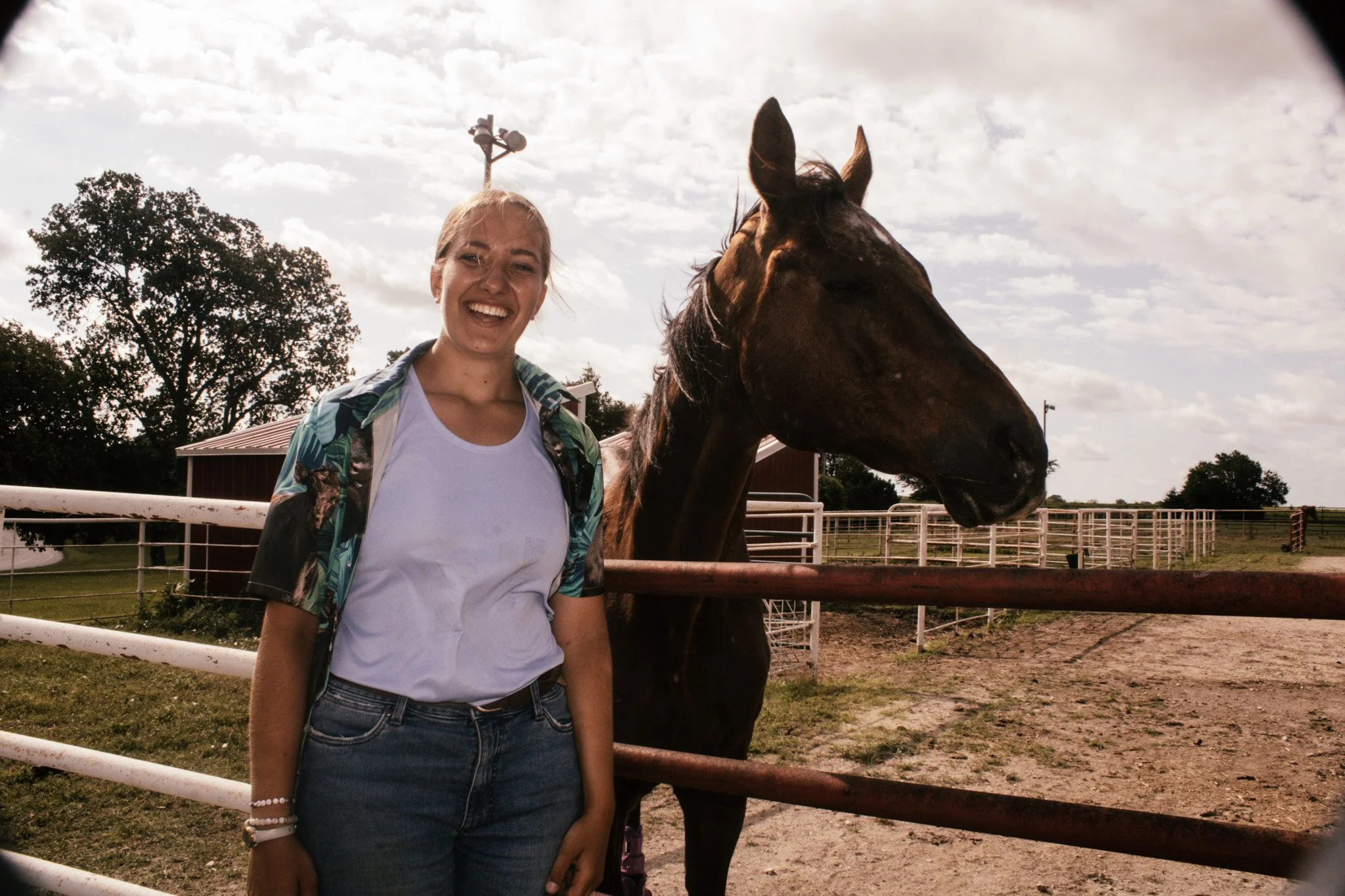 A young woman standing in front of a brown horse at a farm or ranch, with a white fence, trees, and cloudy sky in the background.