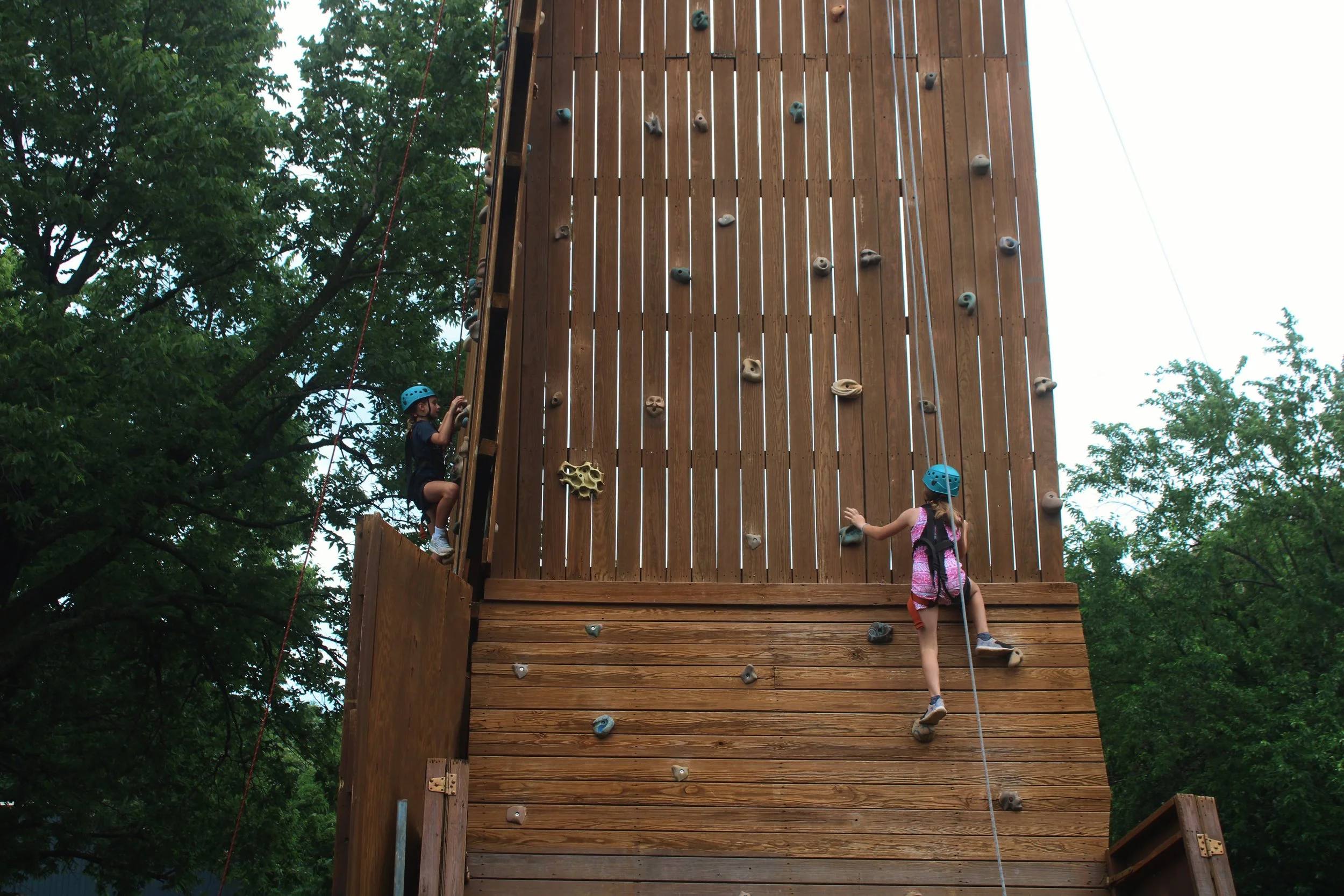 Children climbing a large outdoor wooden rock climbing wall, surrounded by trees.