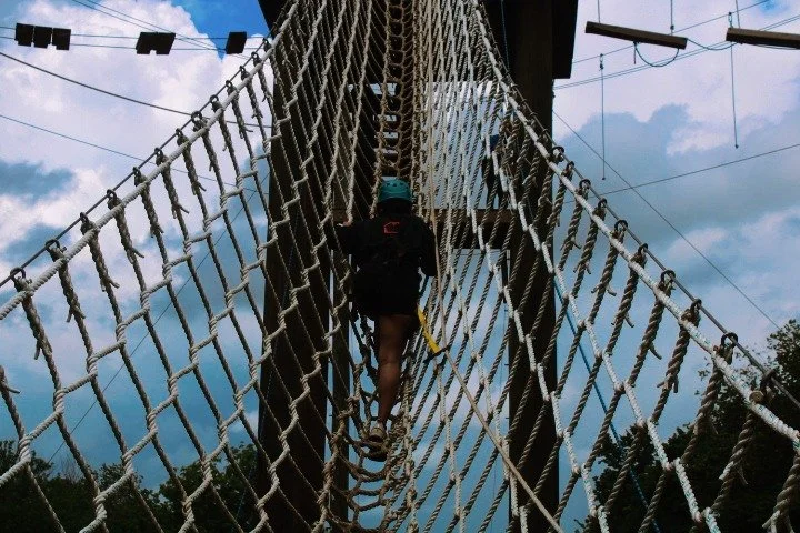 Person wearing a helmet climbing a rope obstacle on a tall outdoor adventure course against a cloudy sky.