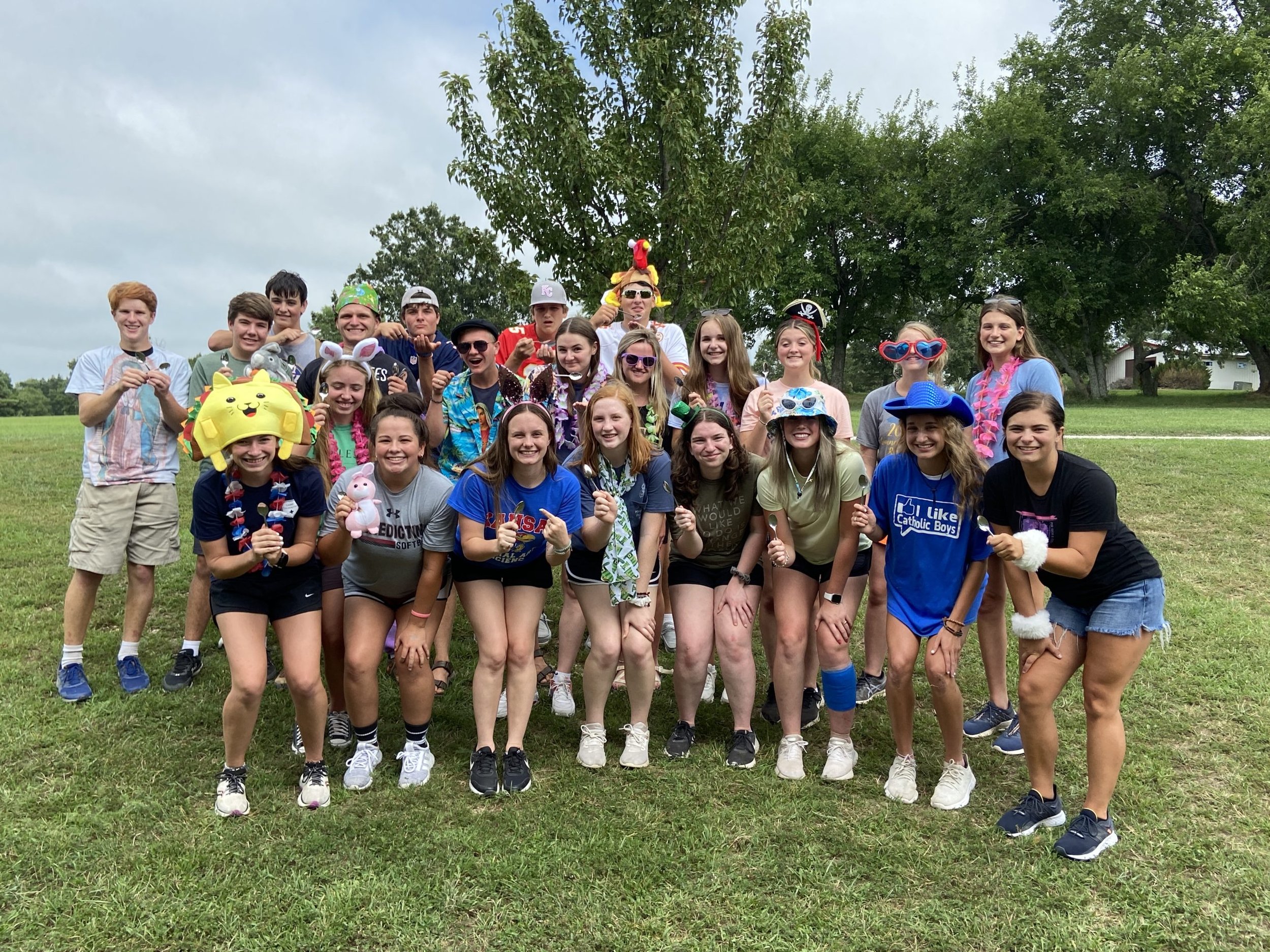 A group of teenagers and young adults standing outdoors on a grassy field, dressed in colorful and playful outfits with accessories like hats, glasses, and headbands, smiling and posing for the camera.