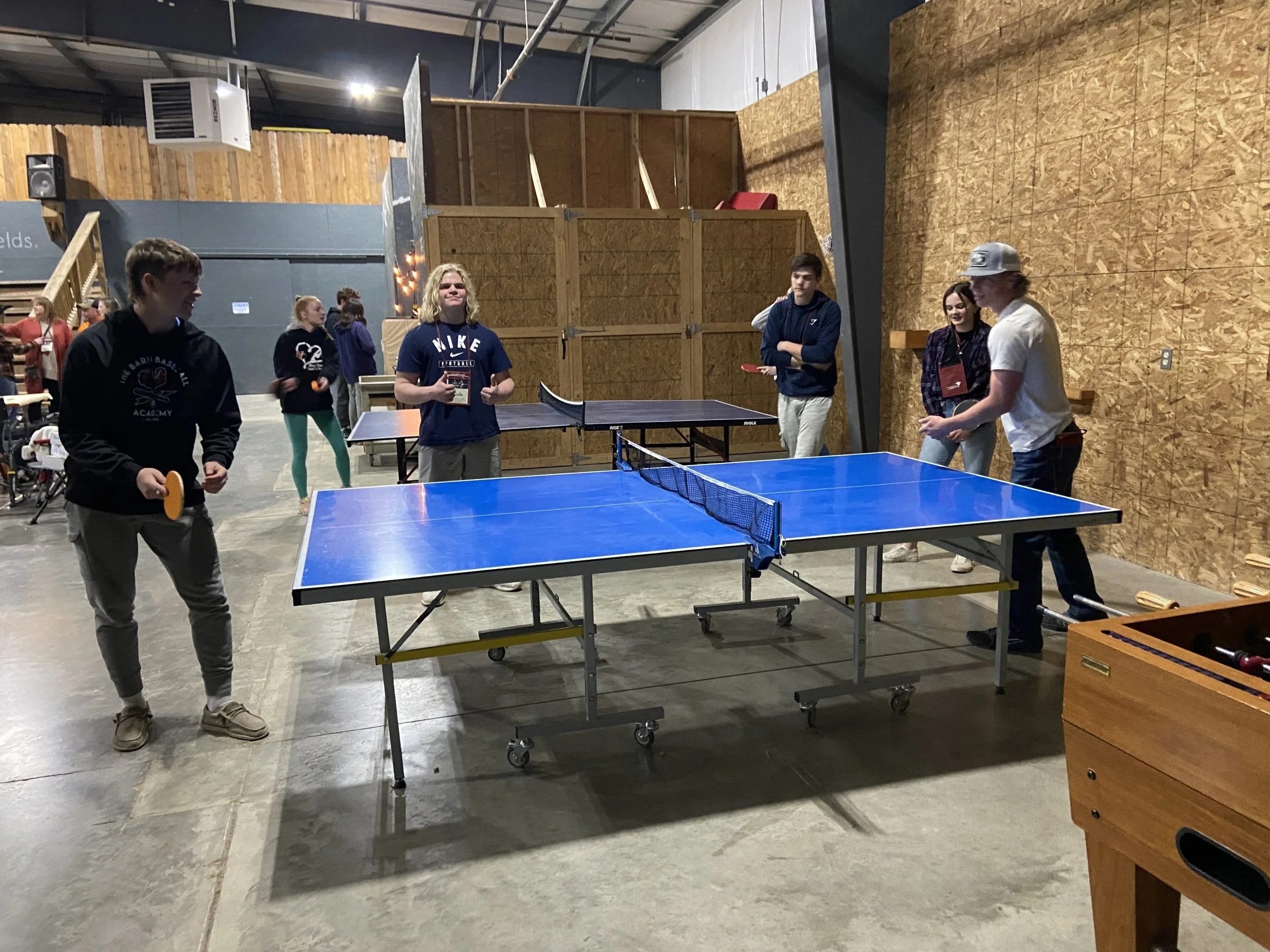 A group of young people playing ping pong indoors, with wooden walls and a concrete floor, in what appears to be a recreational or community space.
