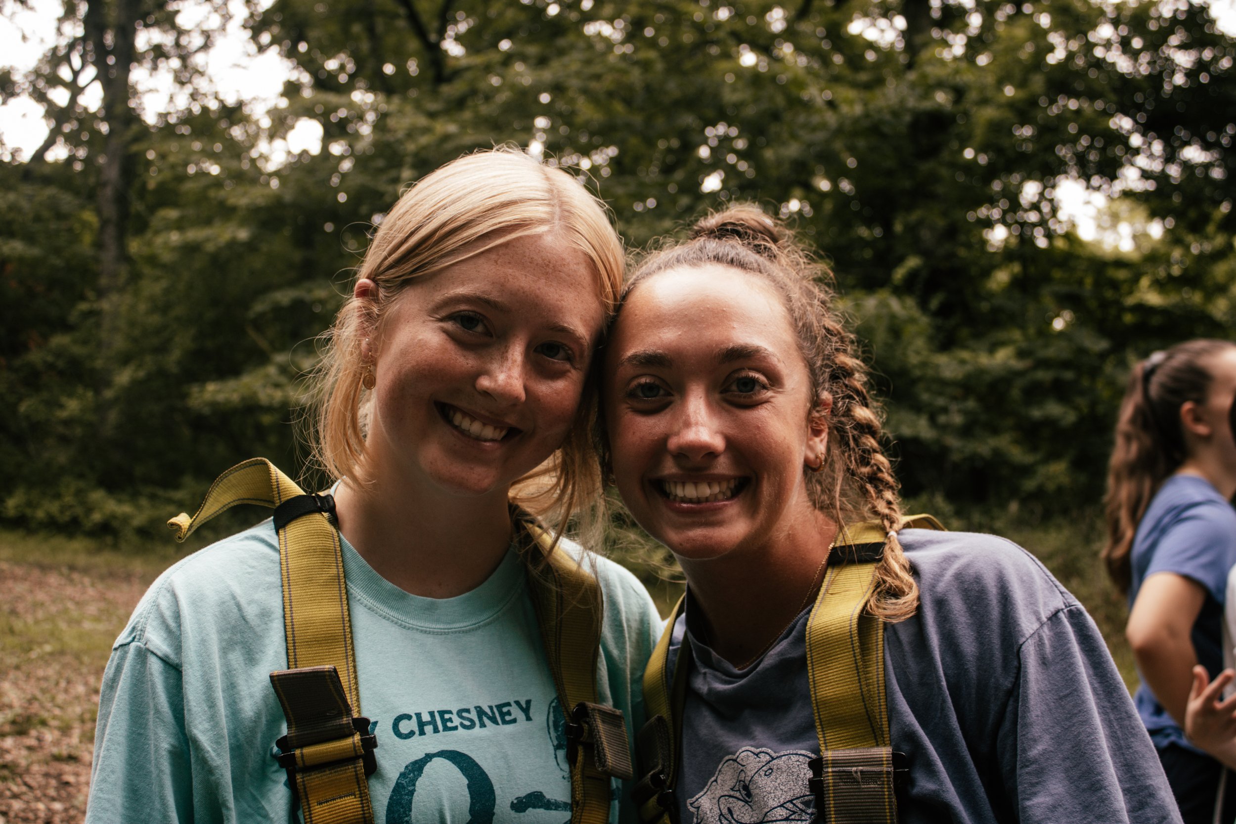 Two young women smiling outdoors during daytime, wearing backpacks, surrounded by trees.