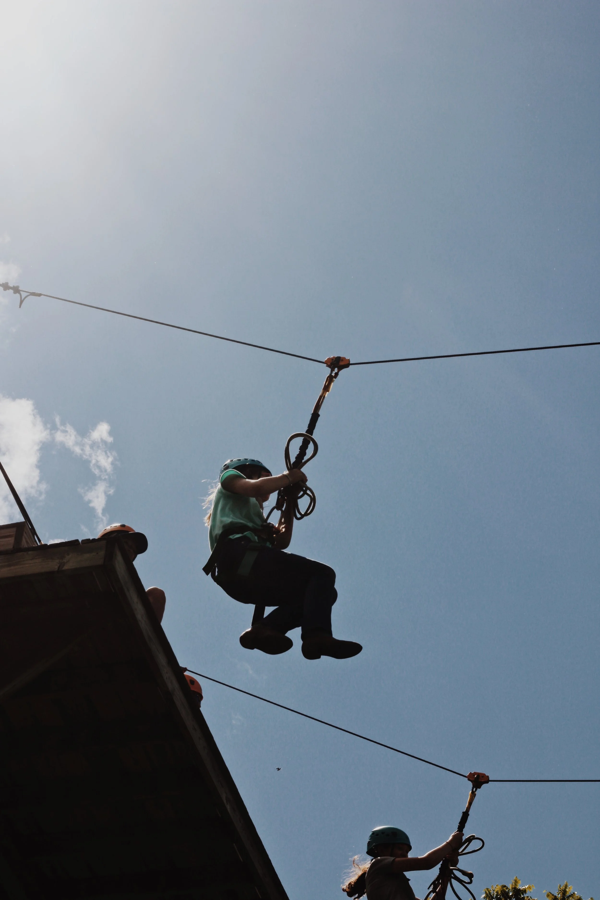 Two children wearing helmets are on a zipline against a blue sky with some clouds. One child is on the lift platform, while the other is holding onto the zipline cable.