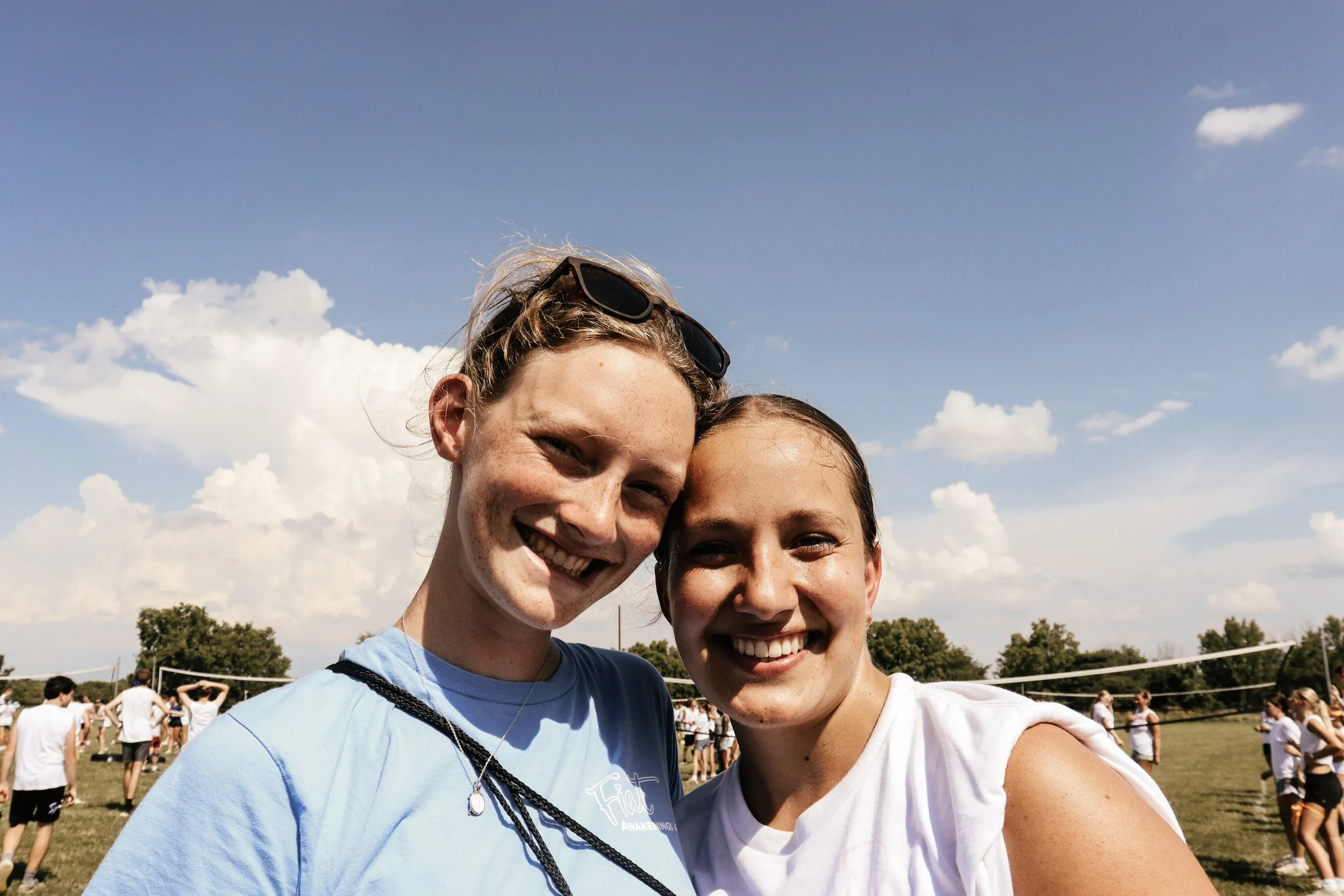 Two young women smiling and taking a selfie outdoors with a large crowd in the background on a sunny day.
