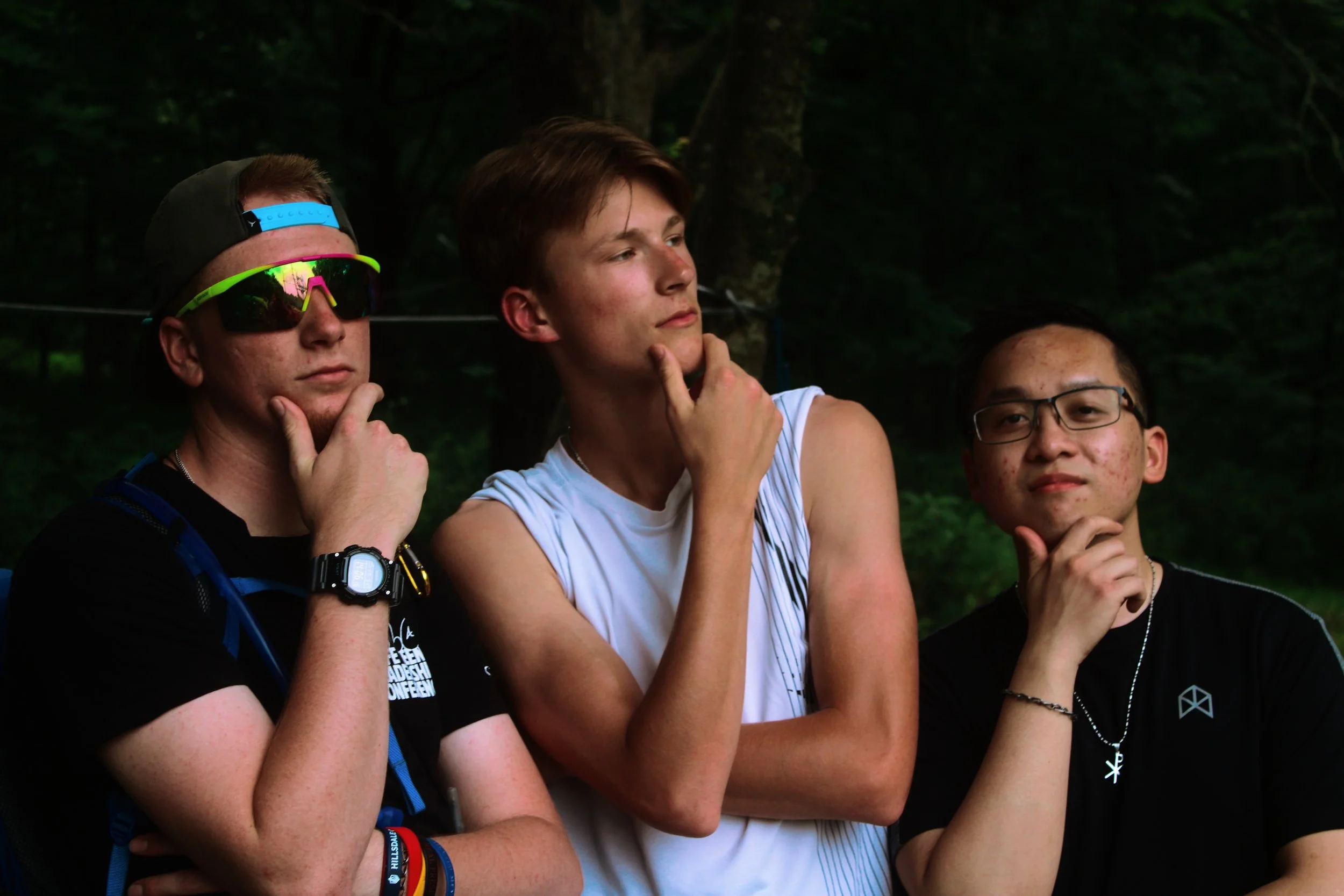 Three young men posing outdoors with thoughtful expressions, three of their hands touching their chins, surrounded by a dark green forest background.
