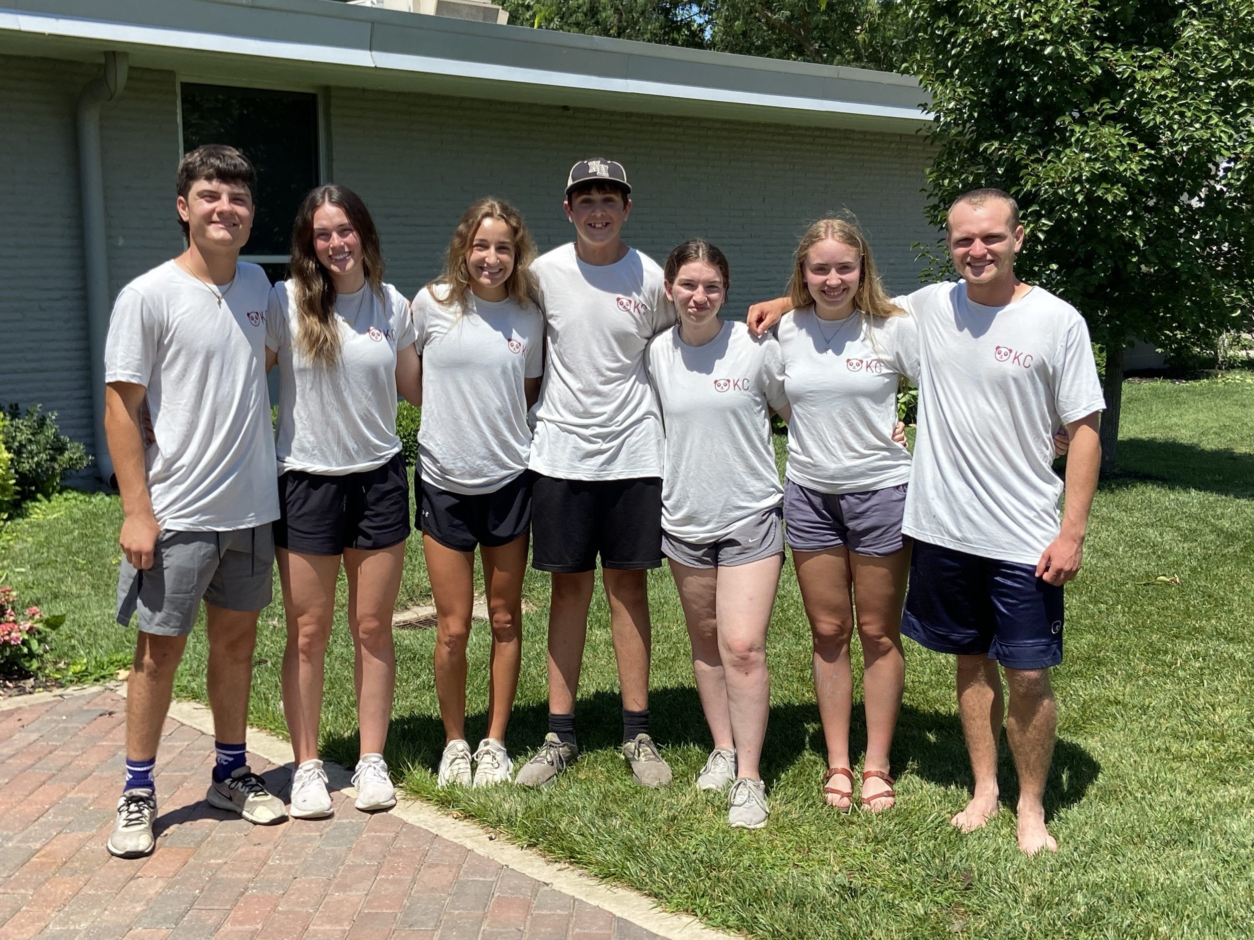 A group of seven young people, six girls and one boy, standing together outside on a sunny day in front of a house with a brick walkway and a green lawn. They are all wearing white T-shirts with a pink and purple logo on the front and are smiling at 