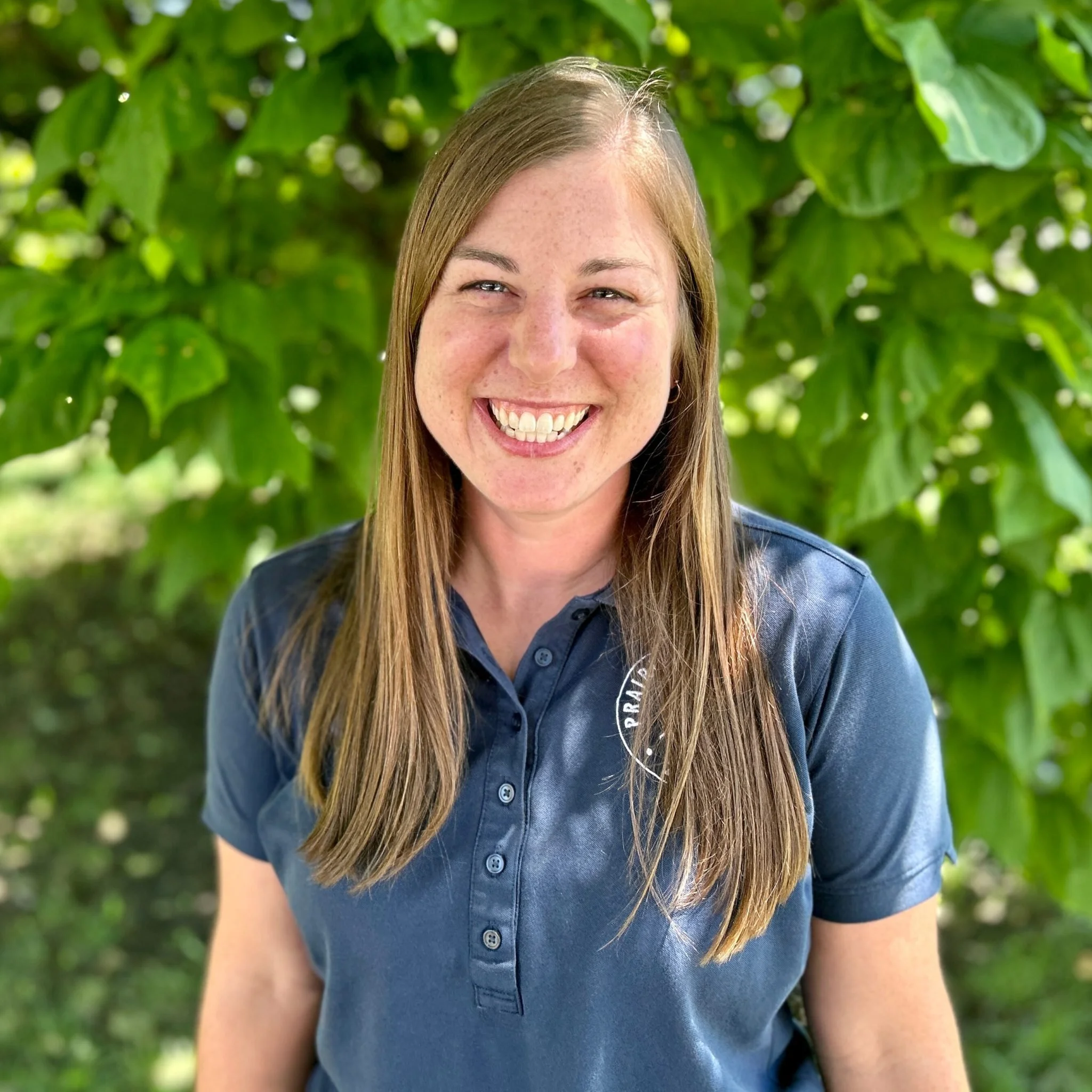 Close-up of a woman with long brown hair smiling, wearing a navy blue collared shirt, standing outdoors with green foliage in the background.