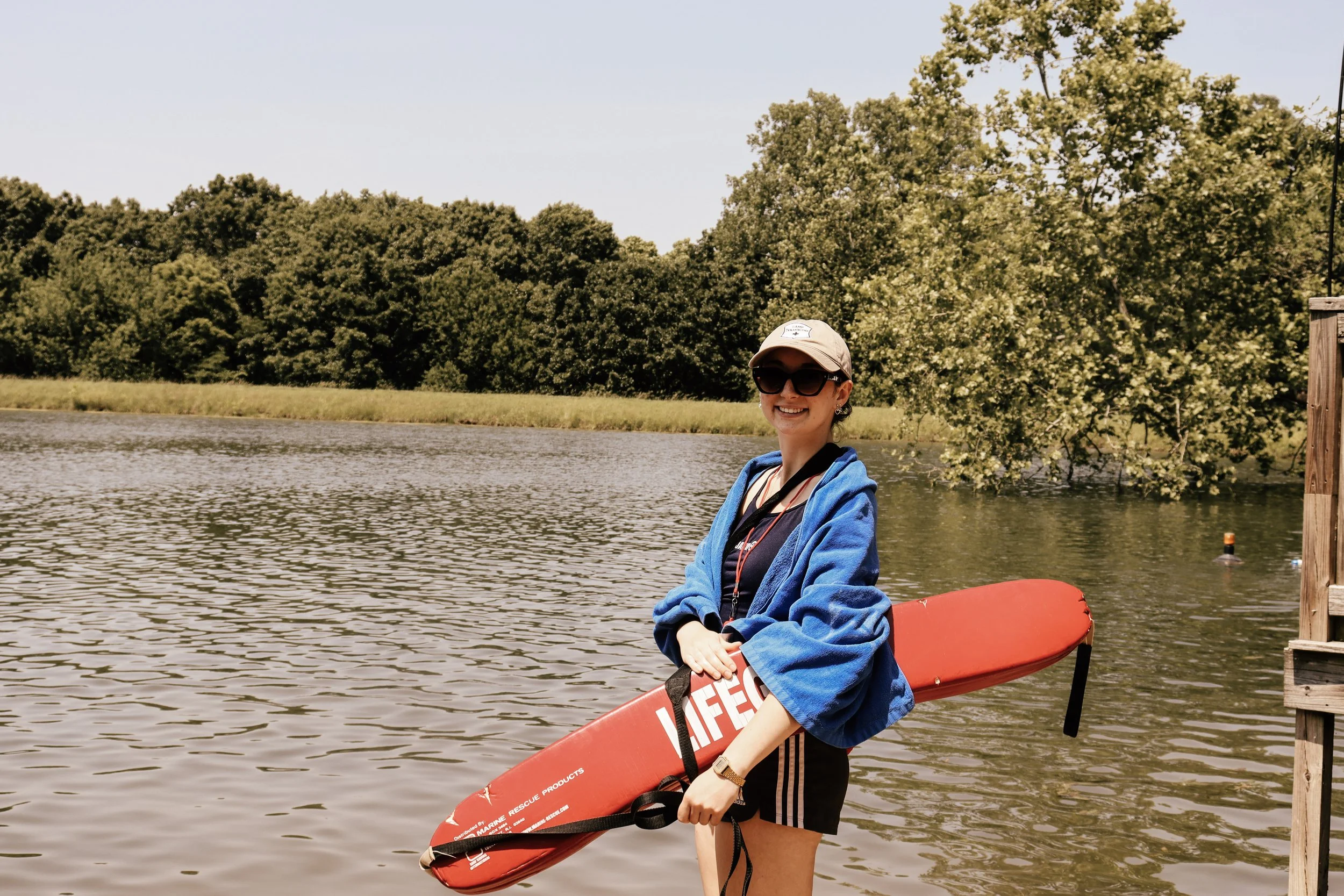 A woman holding a life rescue board while standing near a river, wearing sunglasses, a baseball cap, a blue hoodie, and shorts, with trees and a wooden dock in the background.