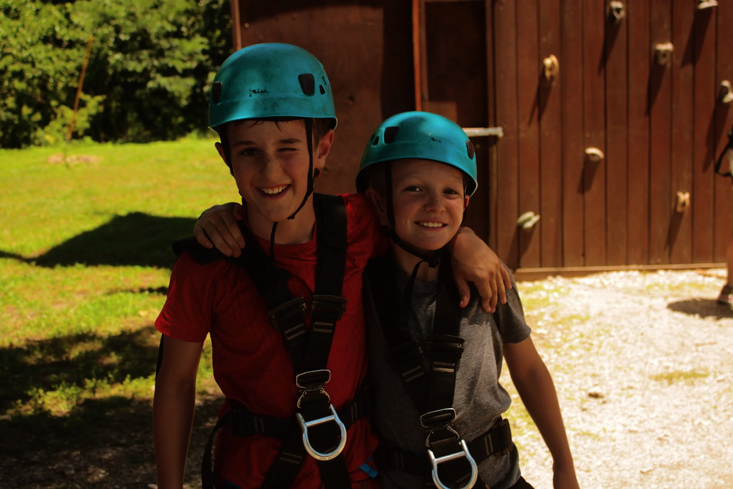 Two children wearing blue helmets and harnesses smiling outdoors in front of a wooden climbing wall with greenery in the background.