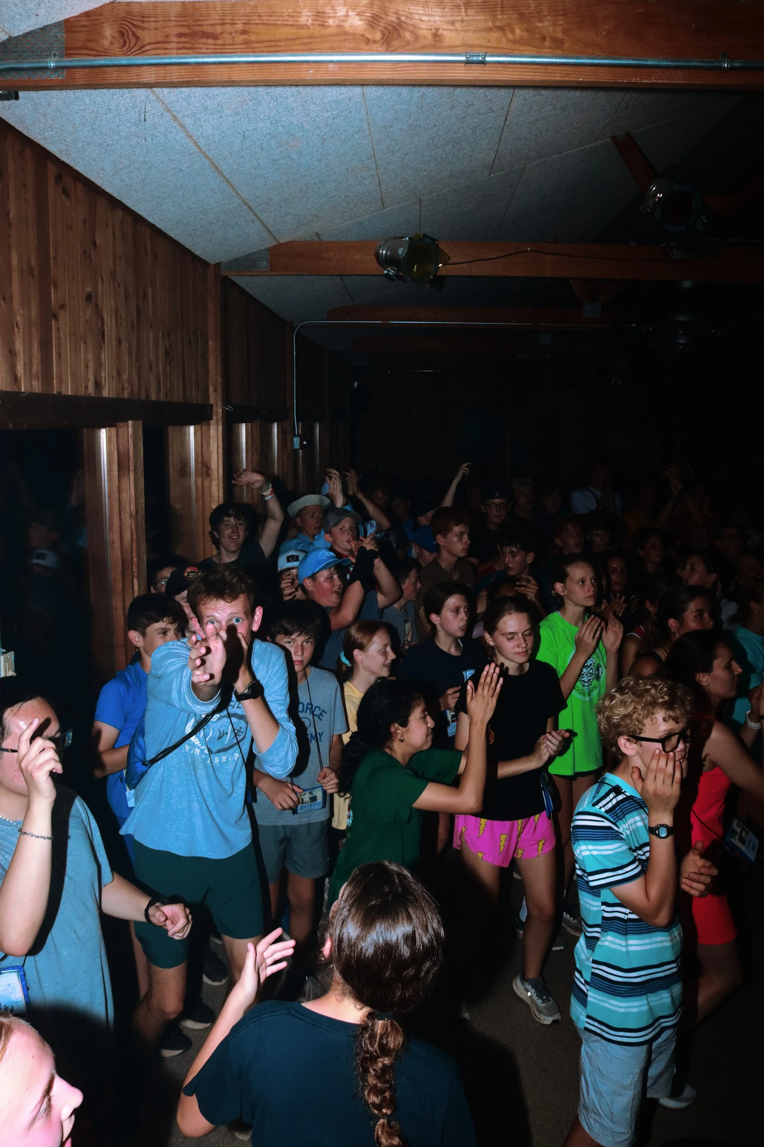 A large group of young people gathered in a dimly lit room with wooden paneling, some raising their hands and clapping, with a few appearing to sing or shout.