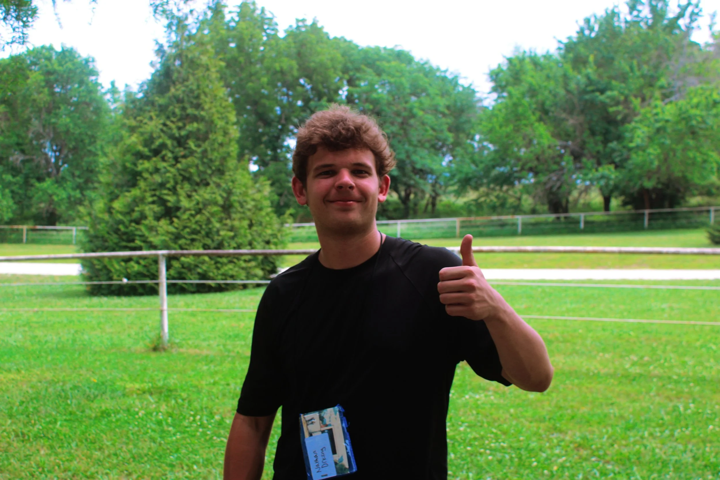 Young man with curly hair smiling and giving a thumbs-up, standing on green grass with trees in the background.