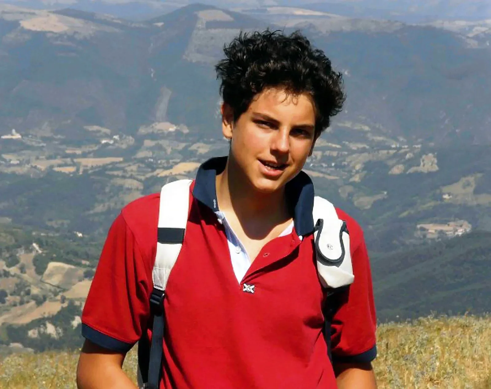 Young man hiking in mountainous area, wearing a red shirt and carrying a white backpack.
