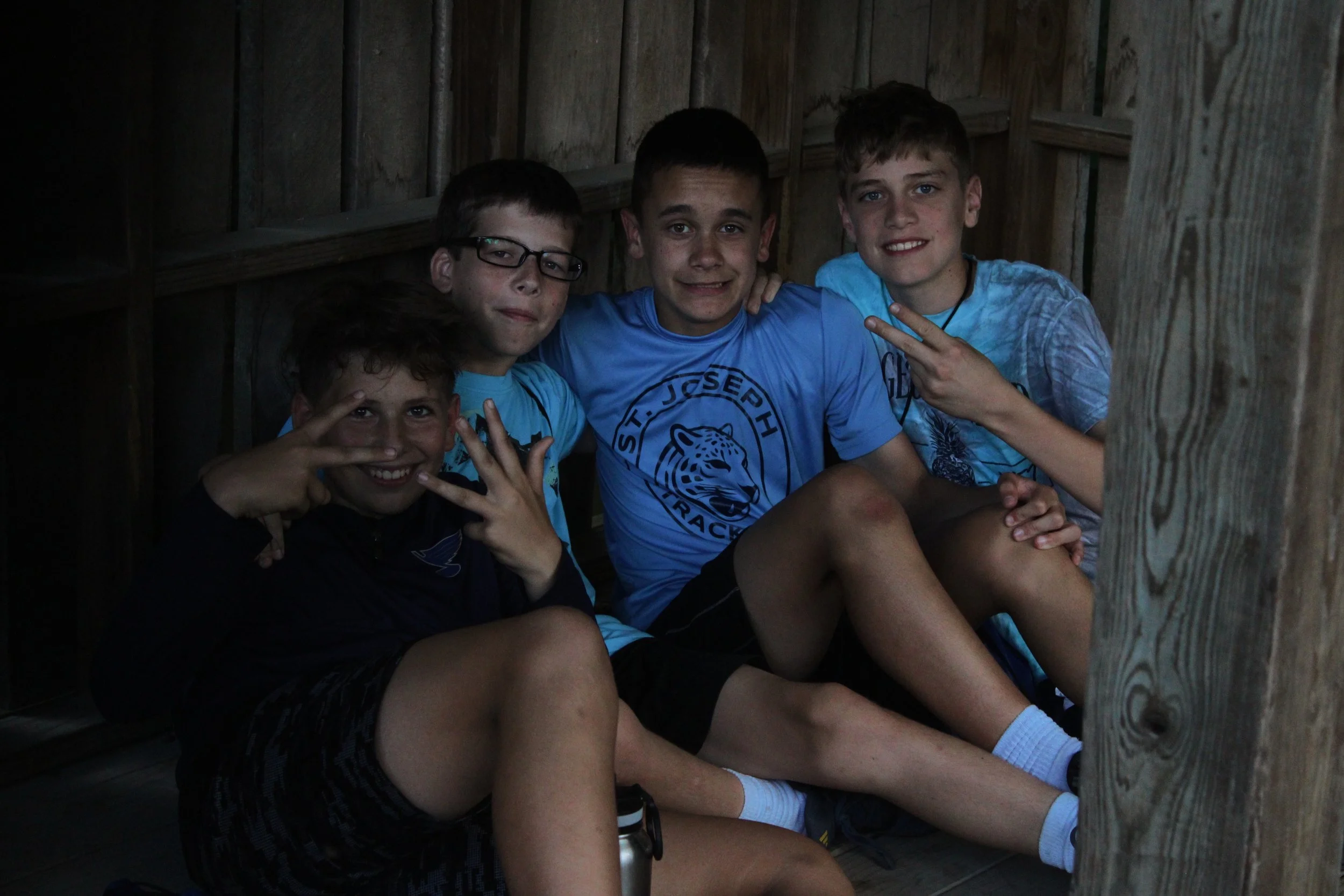 Four boys sitting inside a wooden structure. They are smiling and making peace signs with their hands.