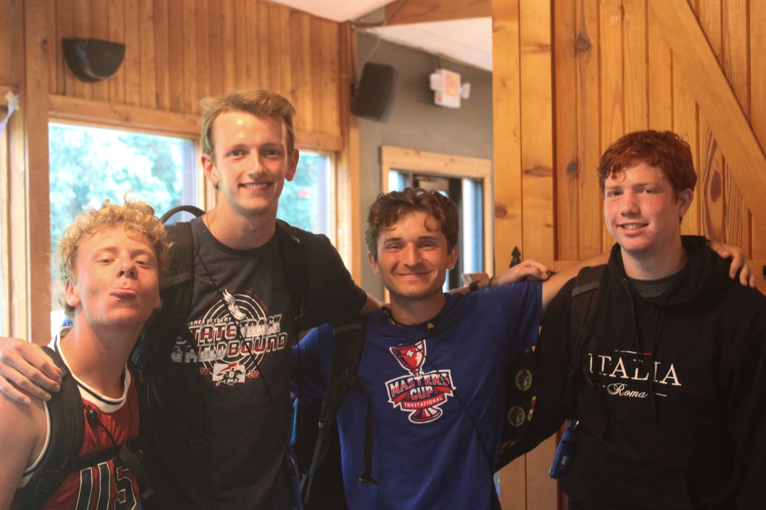 Group of four young men with backpacks indoors, smiling and posing for a photo, wooden walls in the background.