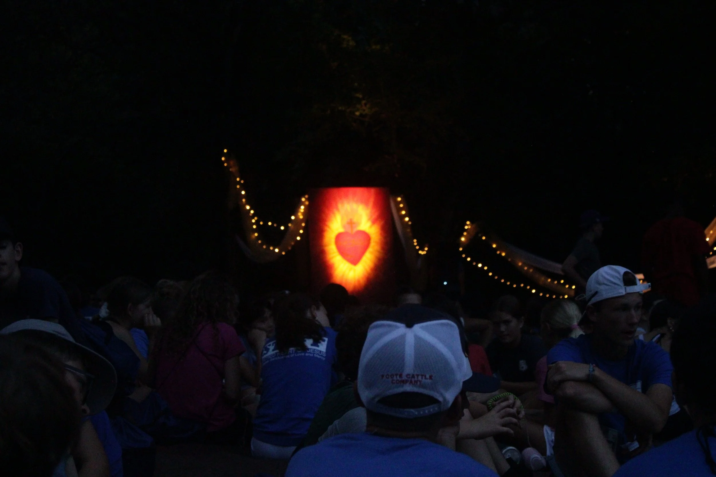 A large crowd of people gathered outdoors at night, with some wearing caps and jackets. A prominent illuminated religious image of a Sacred Heart with a cross above it is displayed in the background, decorated with string lights.