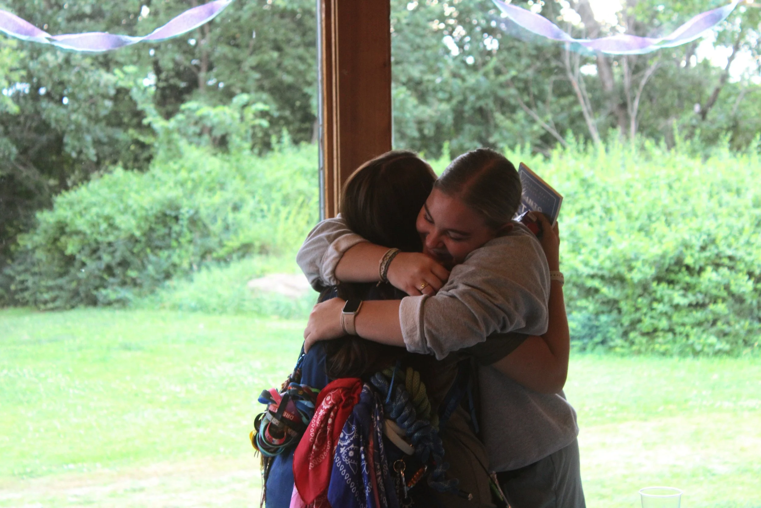 Two people hugging outdoors, likely in a backyard or garden, with trees and greenery visible through a large window in the background.