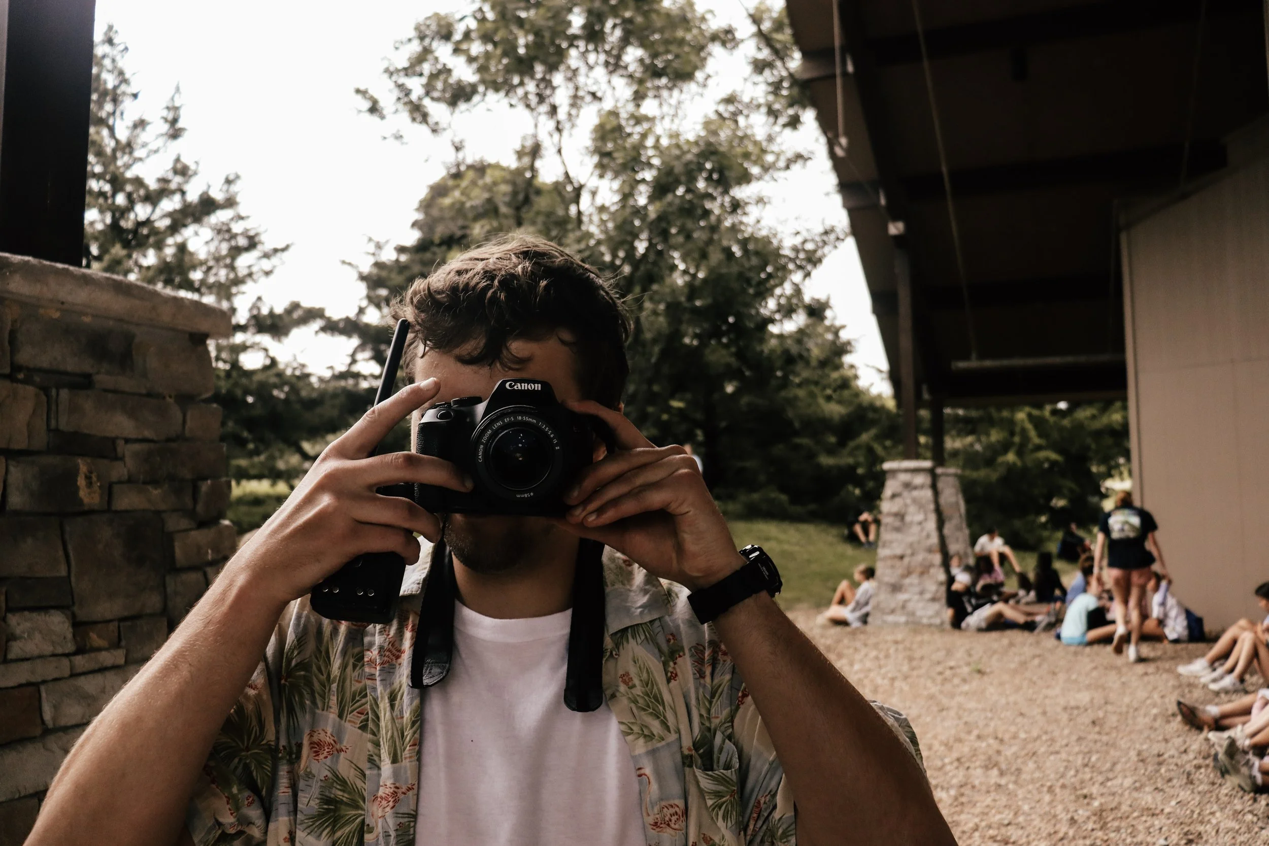 A person takes a photo with a Canon camera outdoors at what appears to be a park or camp site, with other people sitting on ground in the background.