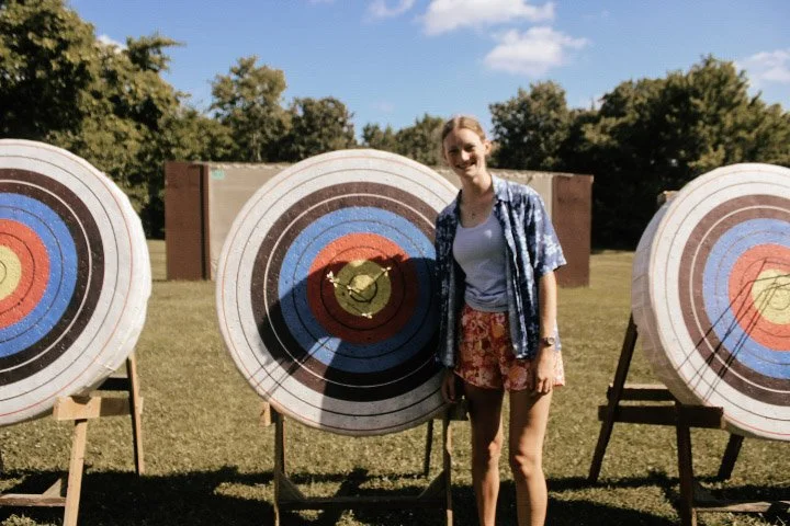 A woman stands outdoors on a sunny day, smiling, between two archery targets with colorful concentric circles, in a grassy field with trees in the background.
