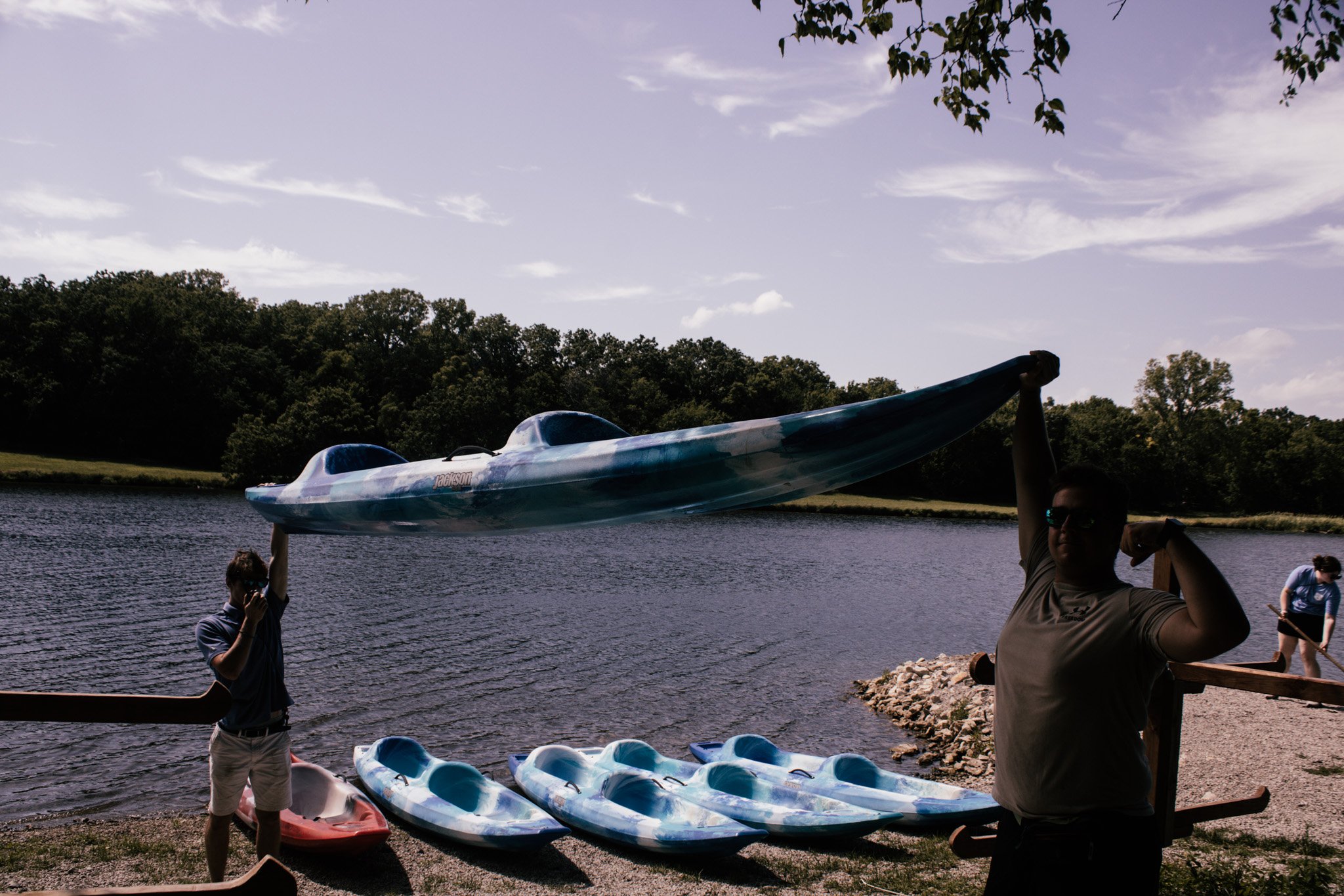 People near a lake holding a kayak on a sunny day, with kayaks on the ground and trees in the background.