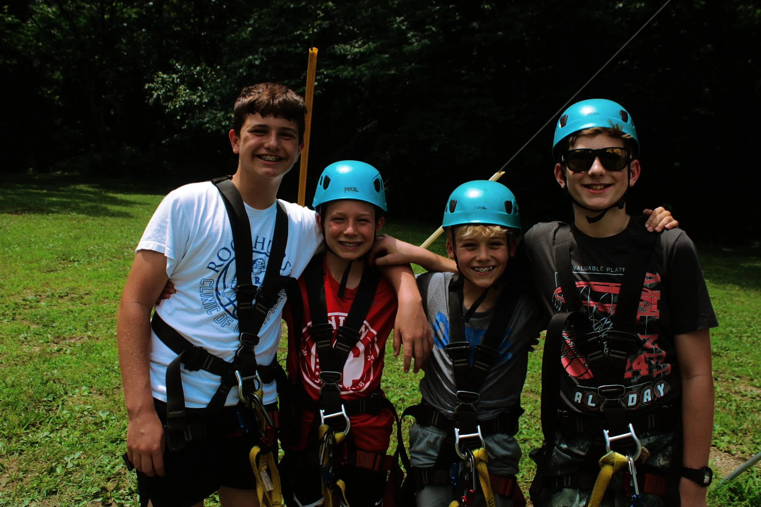 Four boys wearing helmets and harnesses standing outdoors on grass with trees in the background.