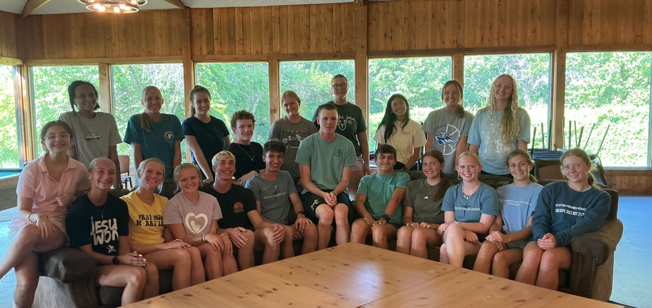 A group of young people, mostly teenagers, sitting and standing in a wooden room with large windows showing green trees outside. They are smiling and appear happy.