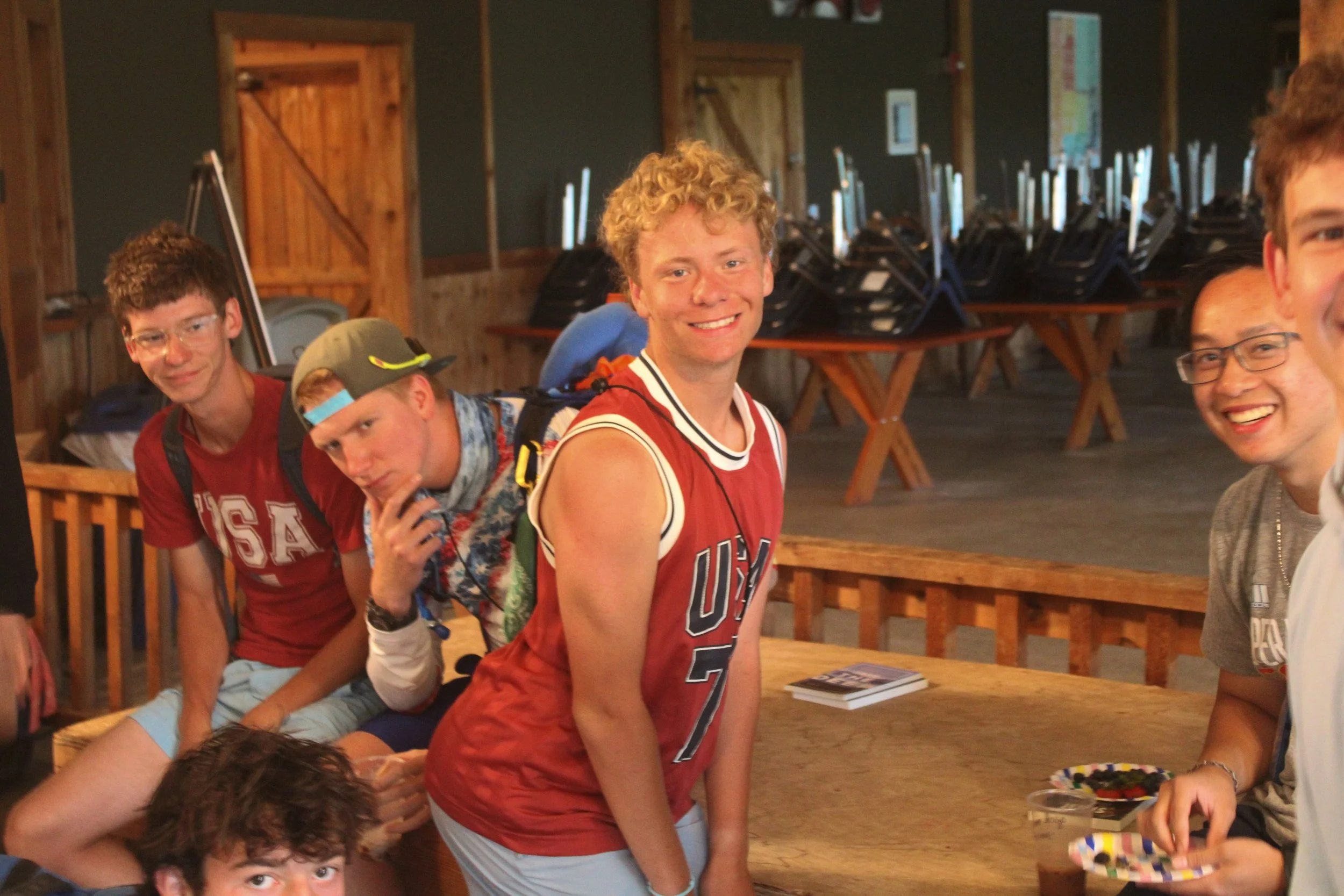 A group of young men smiling and posing at a wooden table in a rustic, wood-paneled room with stacked chairs in the background.