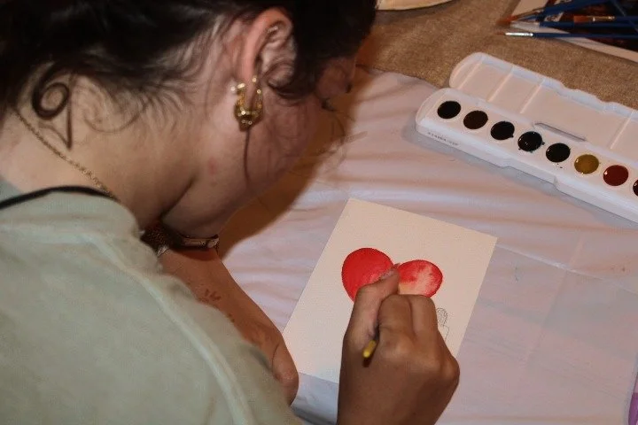 A young woman painting a red heart shape on paper with watercolor paints.