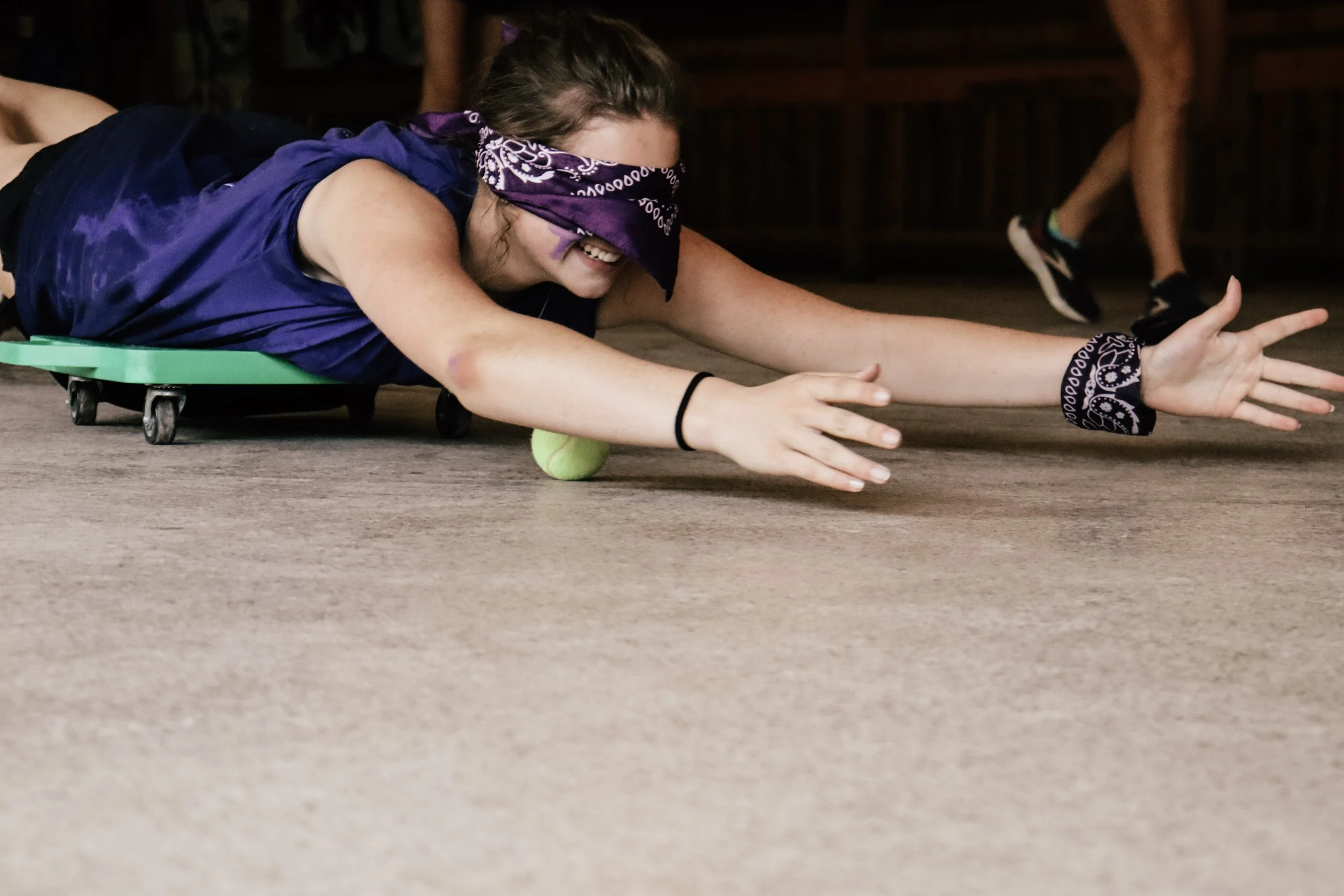 Girl in a purple shirt and bandana blindfolded, smiling as she reaches forward with her hands on a wooden floor, participating in a game involving a small green ball.