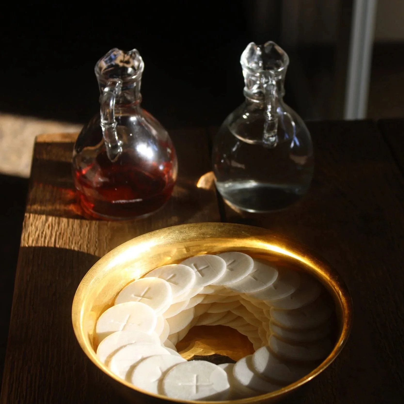 Two glass cruets, one with red liquid and one with clear liquid, on a wooden surface. In front, a golden bowl filled with white round objects, possibly candles or slices of cheese.