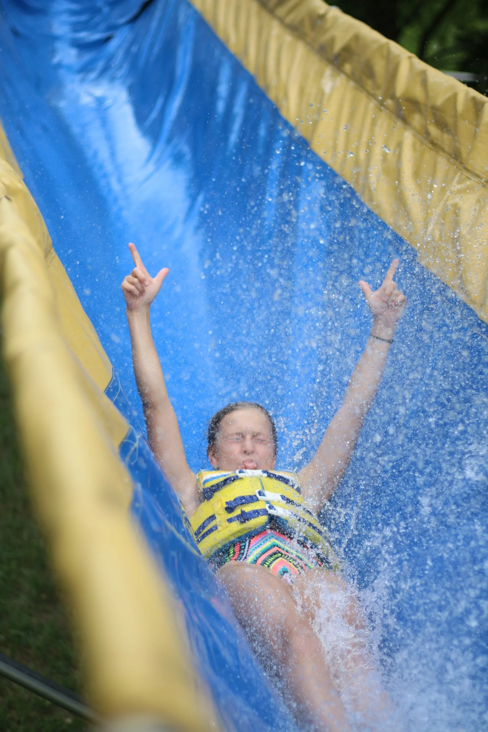 A young girl sliding down a water slide with arms raised, water splashing around her, wearing colorful swimwear, and a life jacket, outdoors on a sunny day.