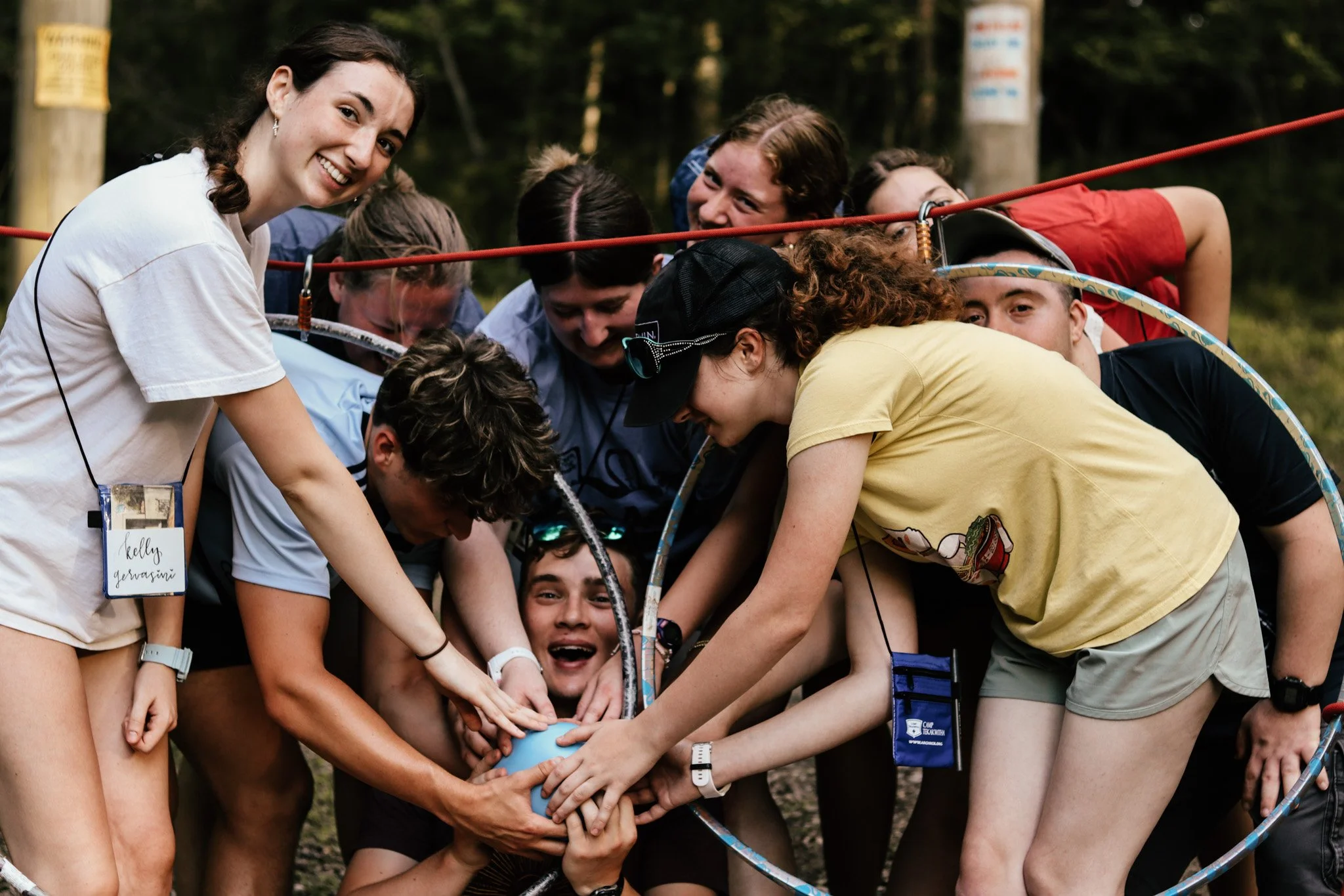 Group of young people participating in an outdoor activity, assembling or launching a small balloon or similar object together, with trees in the background.