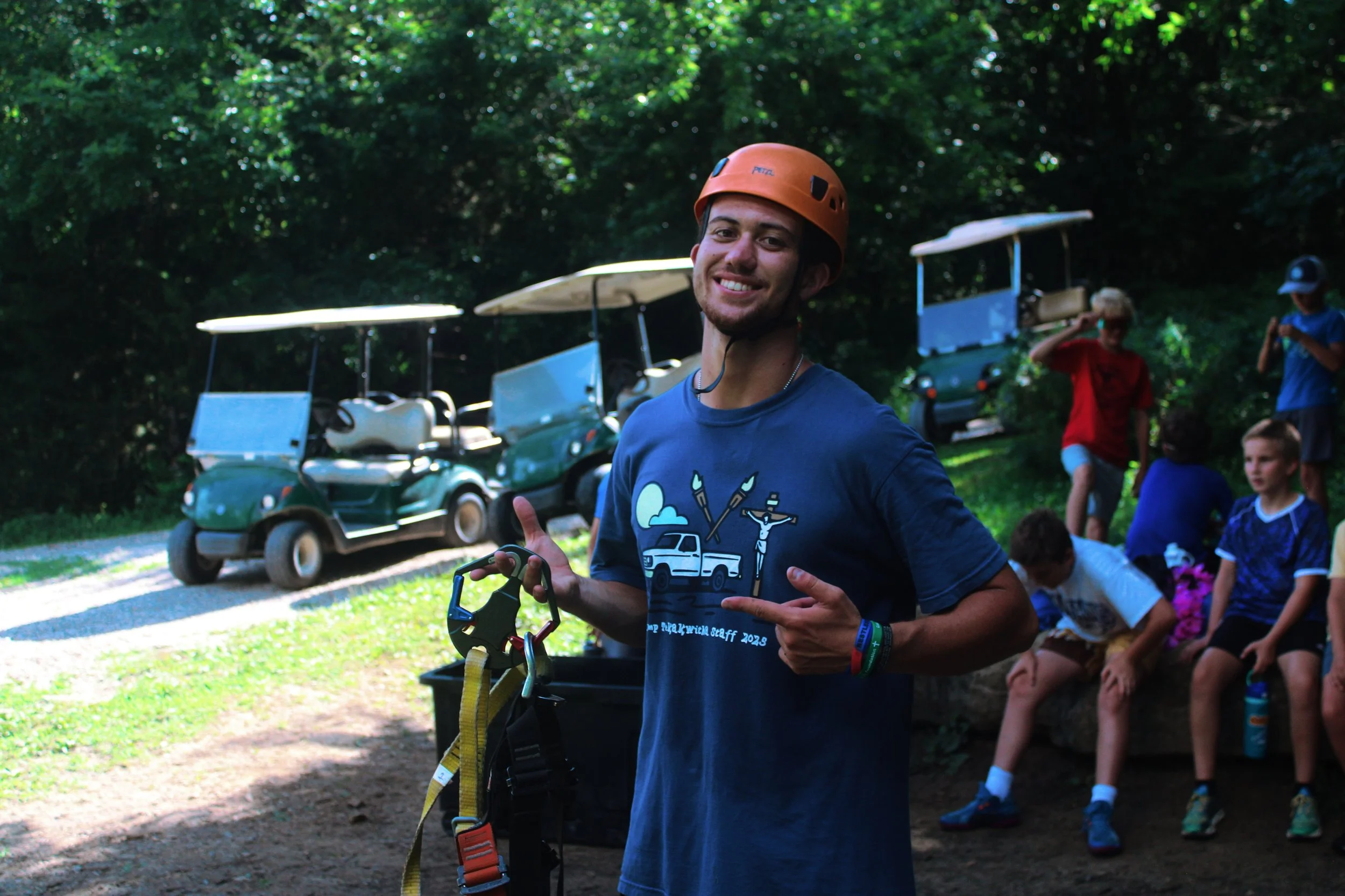 A young man smiling and holding climbing gear outdoors with a group of children sitting on a bench in the background, and golf carts parked among trees.