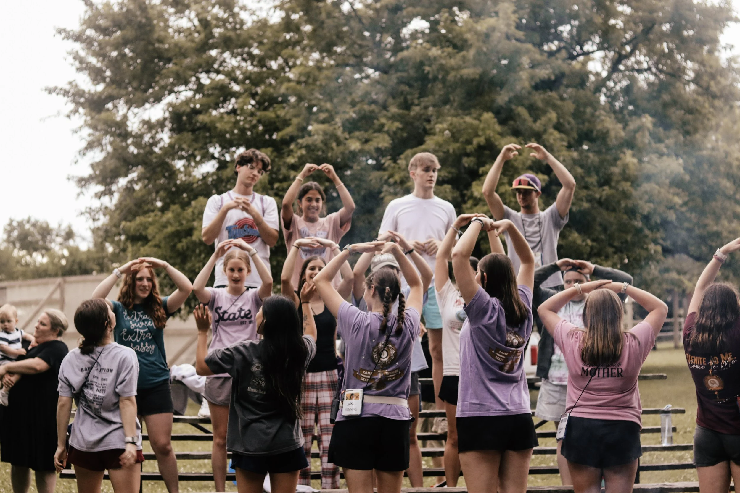 Group of young people gathered outdoors, some forming heart shapes with their hands, with large trees and a cloudy sky in the background.