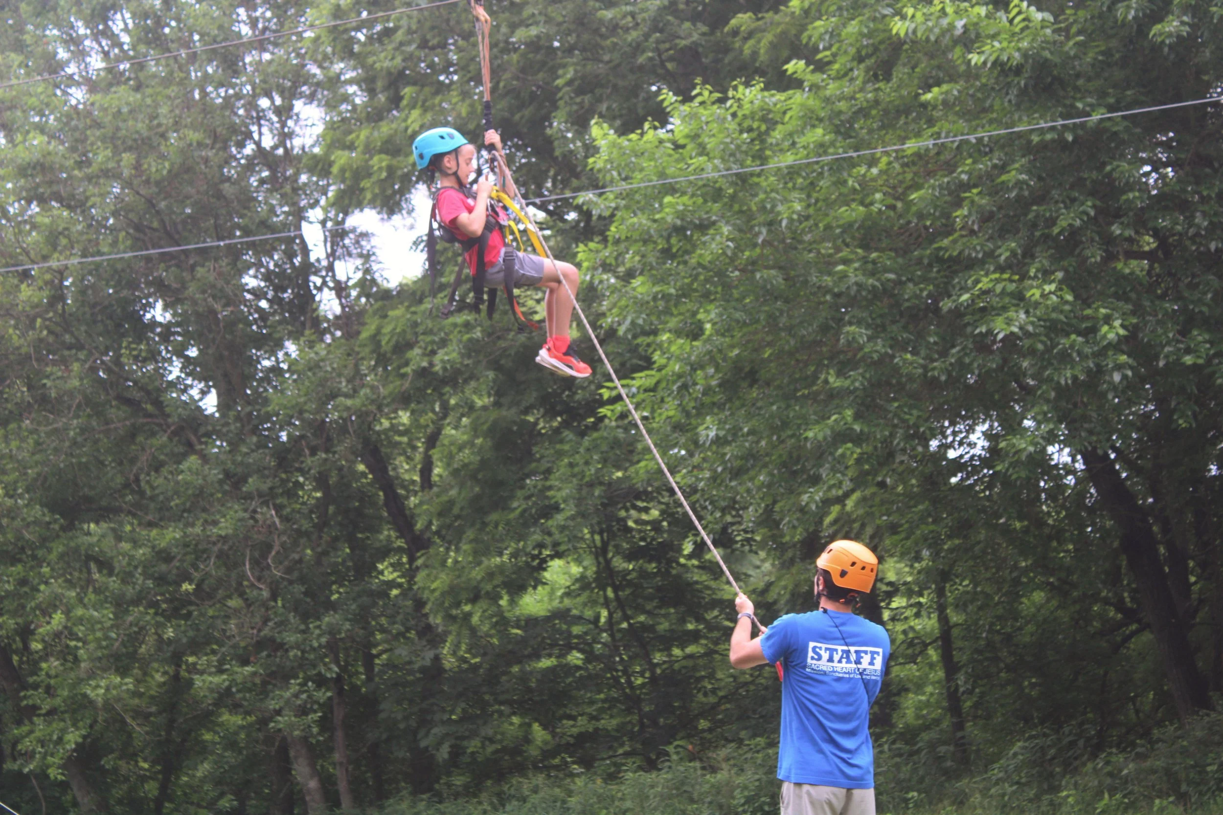 A young boy wearing a blue helmet and pink shirt riding a zip line while an adult man, wearing a yellow helmet and blue staff T-shirt, holds the zip line rope. The background consists of green trees.