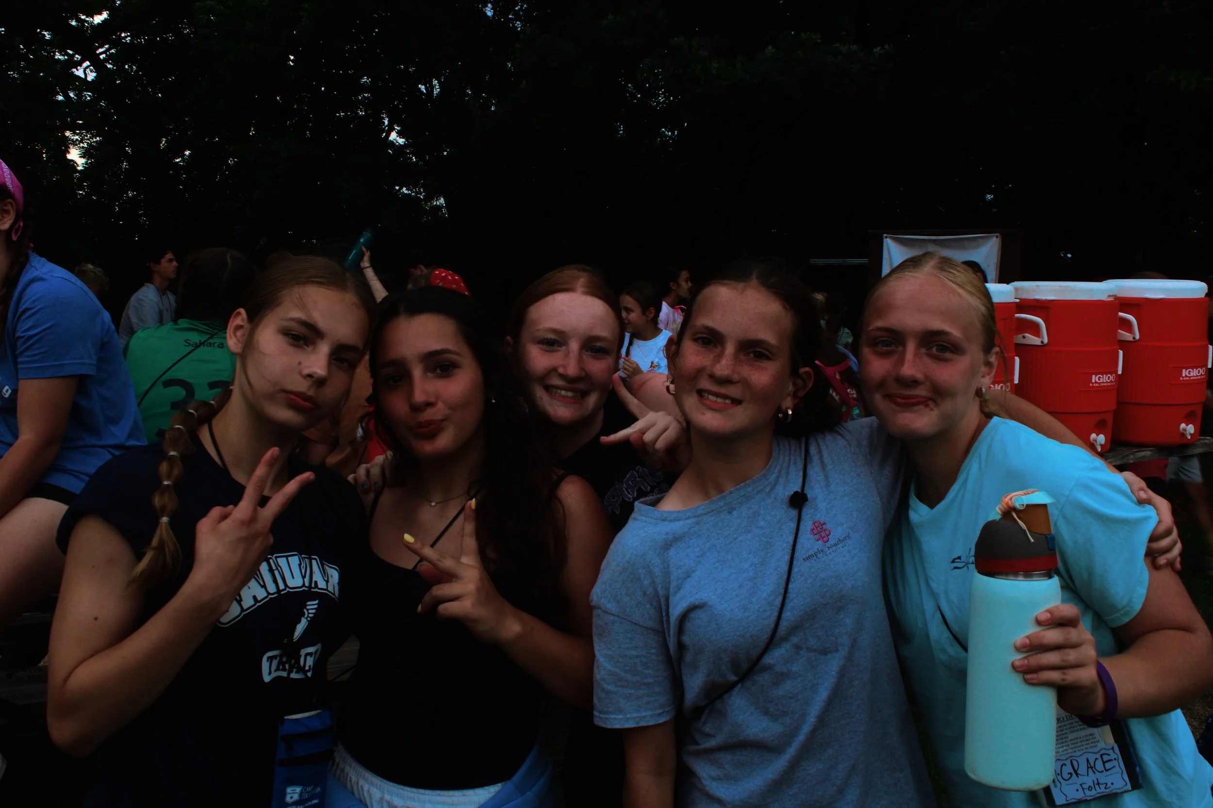 Group of young girls at an outdoor event, smiling and posing for a photo. Some girls are making peace signs and holding water bottles. In the background, there are other people and large orange water coolers, with trees overhead.