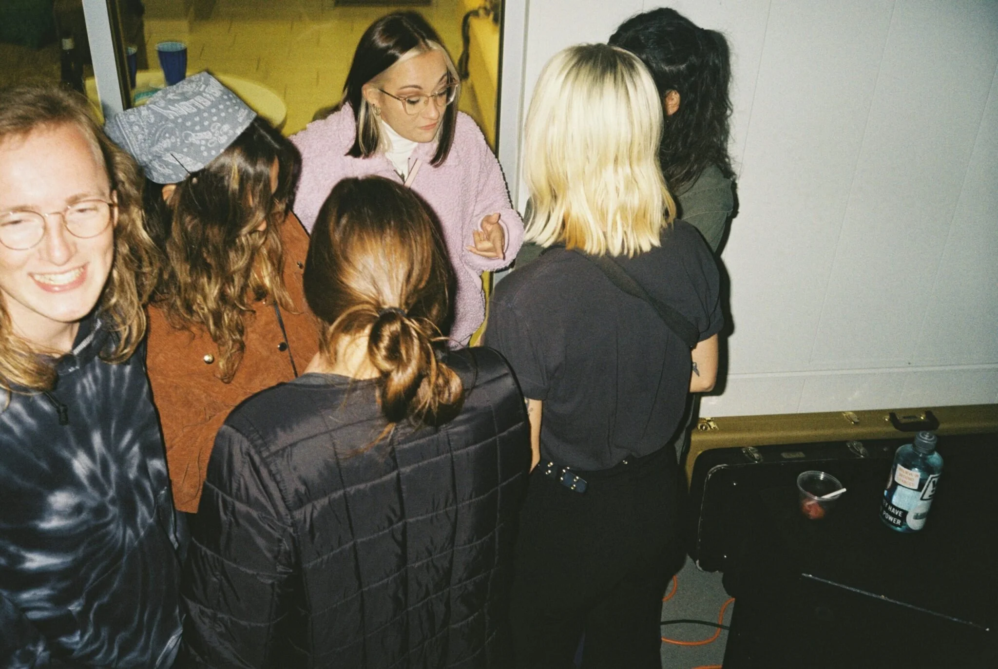 Group of women gathered closely together in an indoor setting, some facing away and others smiling, with beverage cups and bottles on a table nearby.