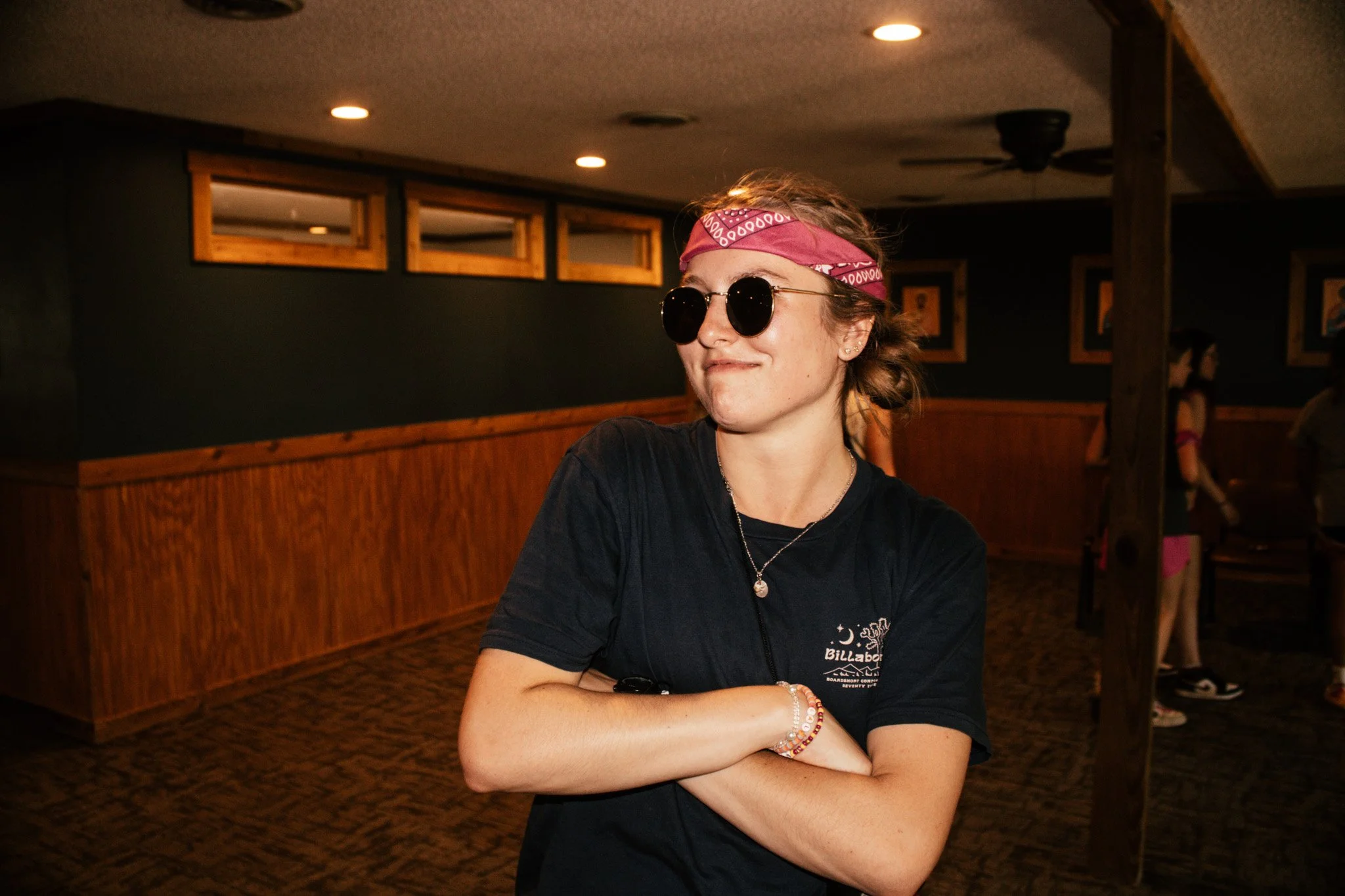 A young woman with wavy hair wearing a pink bandana, round sunglasses, a black t-shirt, and layered bracelets, standing with her arms crossed inside a room with wooden accents and dim lighting.