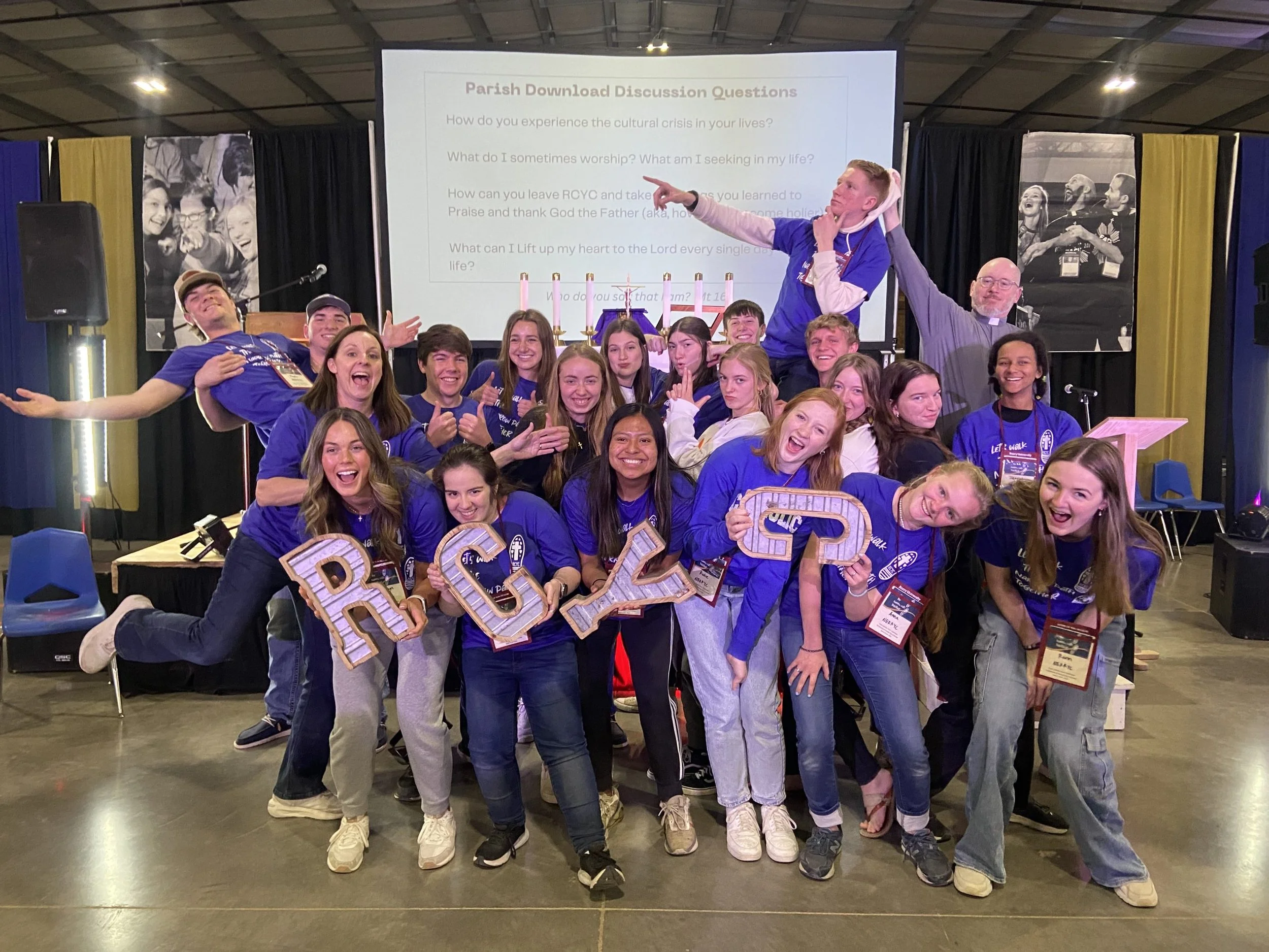 A group of smiling teenagers and a priest posing together at an indoor event, holding large decorated letters spelling B R C 2 3 in the foreground, with a stage, microphones, and large screens displaying discussion questions in the background.
