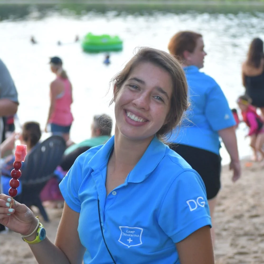 Smiling woman in a blue camp shirt at a lakeside gathering, holding a skewer of food, with other people relaxing by the water.