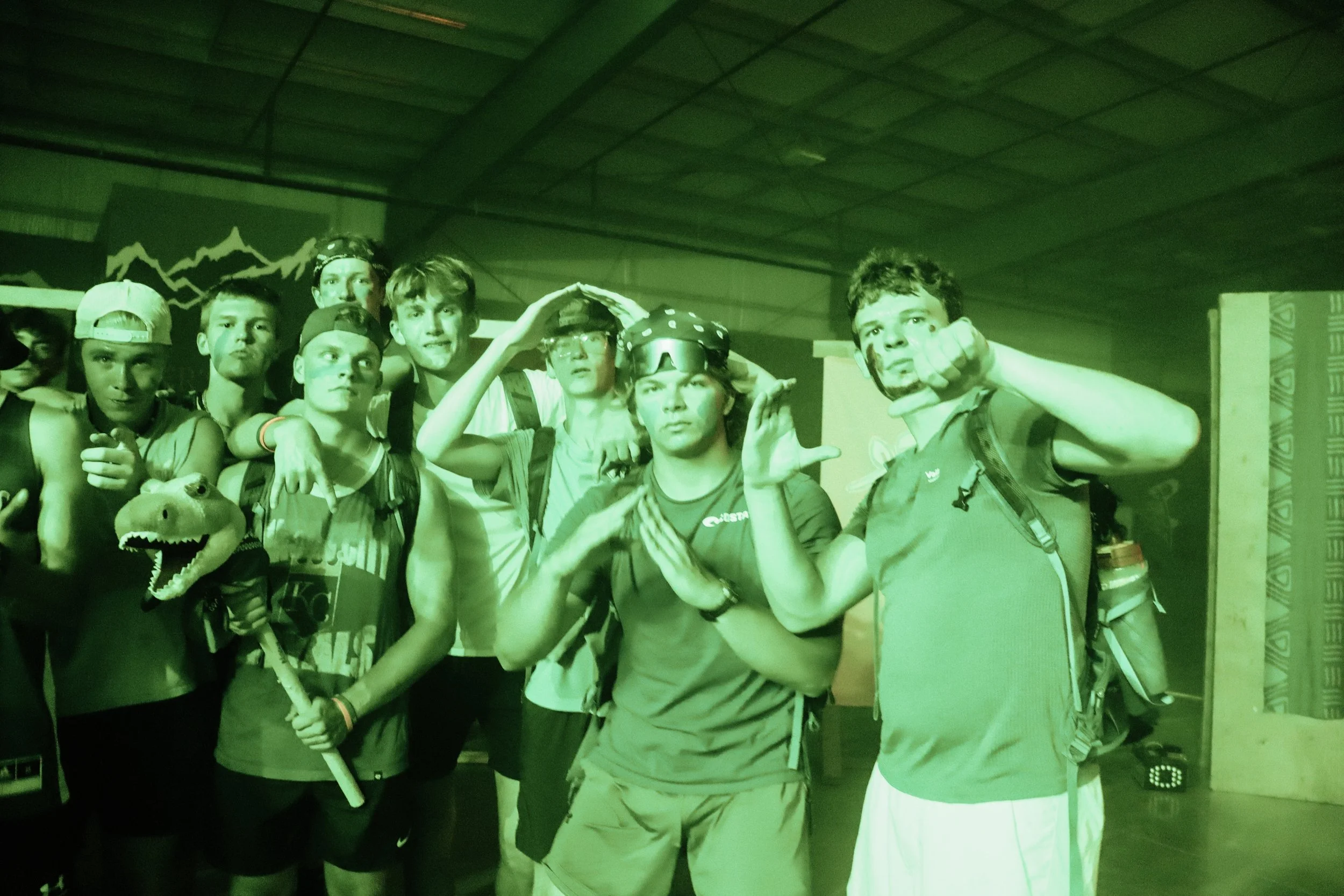 Group of young men dressed in casual and athletic clothing, some with face paint and accessories, posing together in a dimly lit indoor setting with a mountain-themed mural in the background.