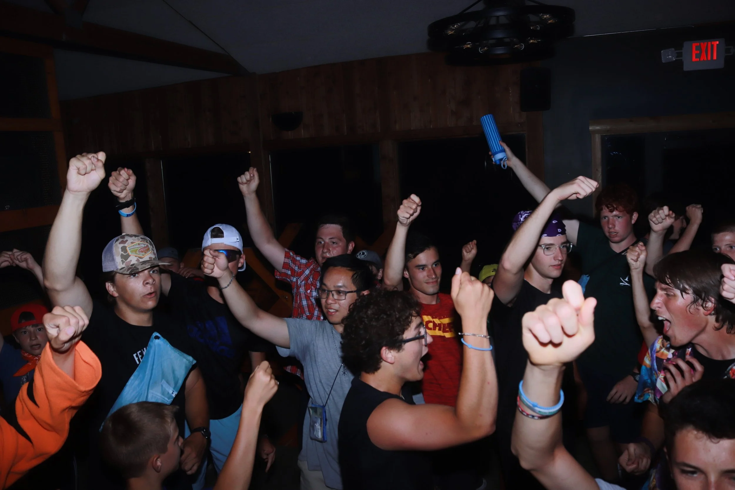 A group of young people celebrating, raising their fists and smiling in a dimly lit indoor space.