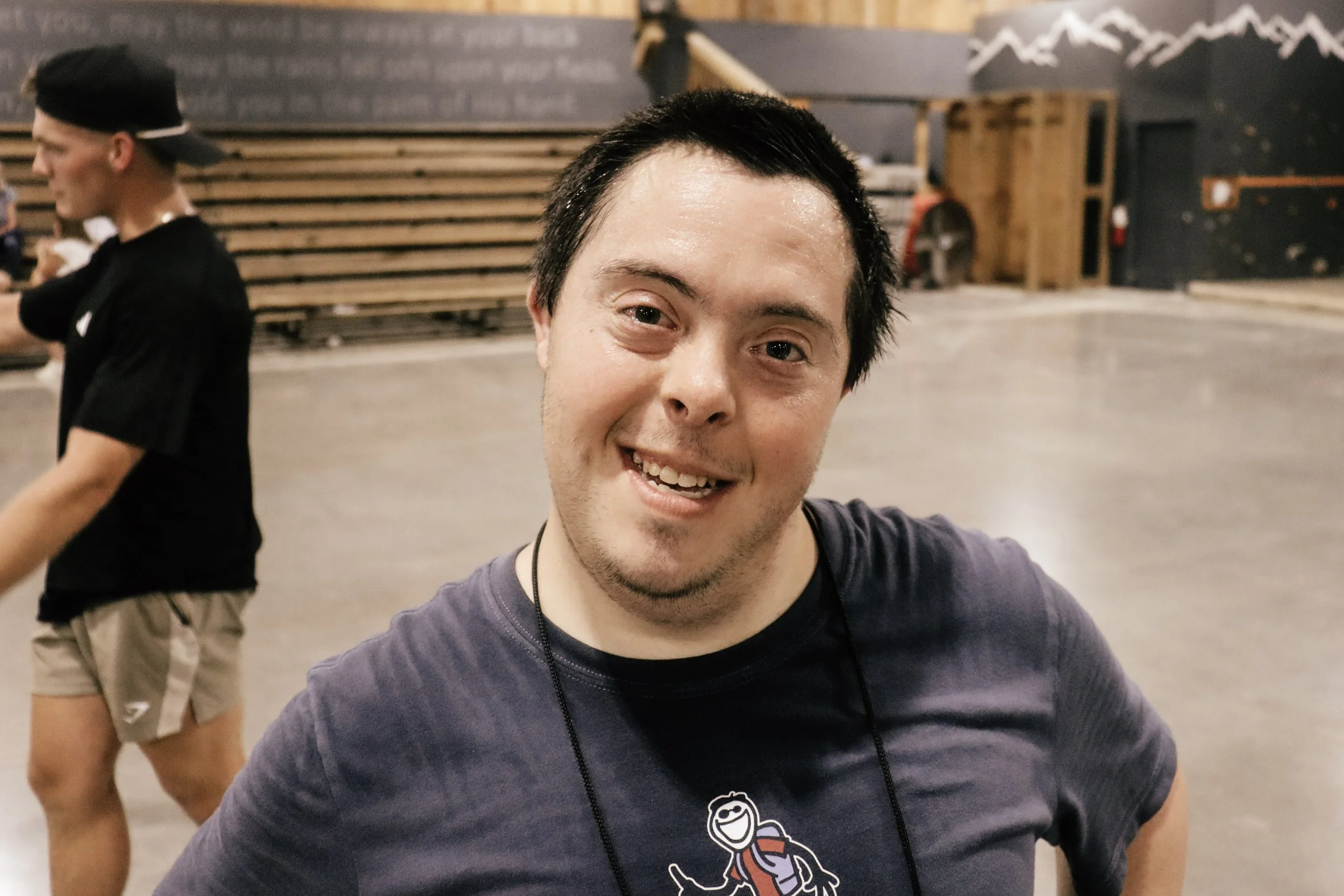 A smiling man taking a selfie with a person wearing a black cap and black shirt in the background. The setting appears to be an indoor space with wooden pallets and a chalkboard with writing and mountains drawn on the wall.