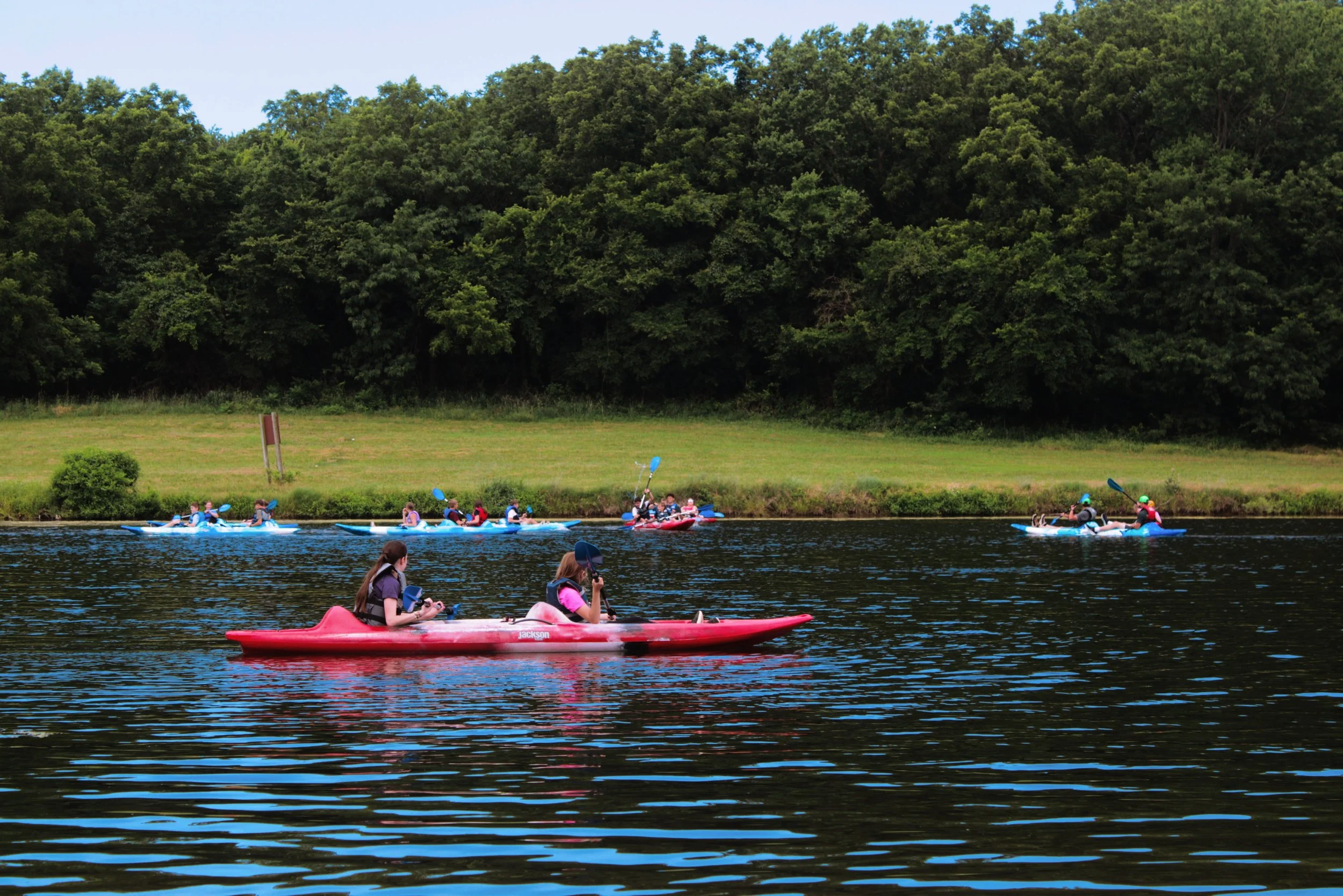 People kayaking on a river with a large green tree-lined hill in the background on a clear day.