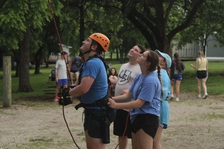 A group of children outside in a park, one girl in a blue shirt and orange helmet is preparing to go rock climbing or rappelling, while others watch and cheer.