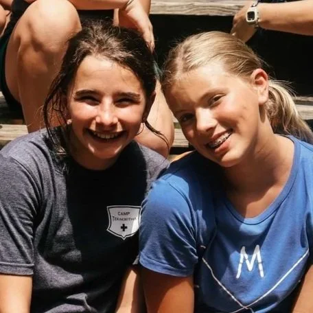 Two young girls smiling outdoors, one wearing a dark gray T-shirt with a camp emblem and the other in a blue T-shirt, with some people's arms visible in the background.